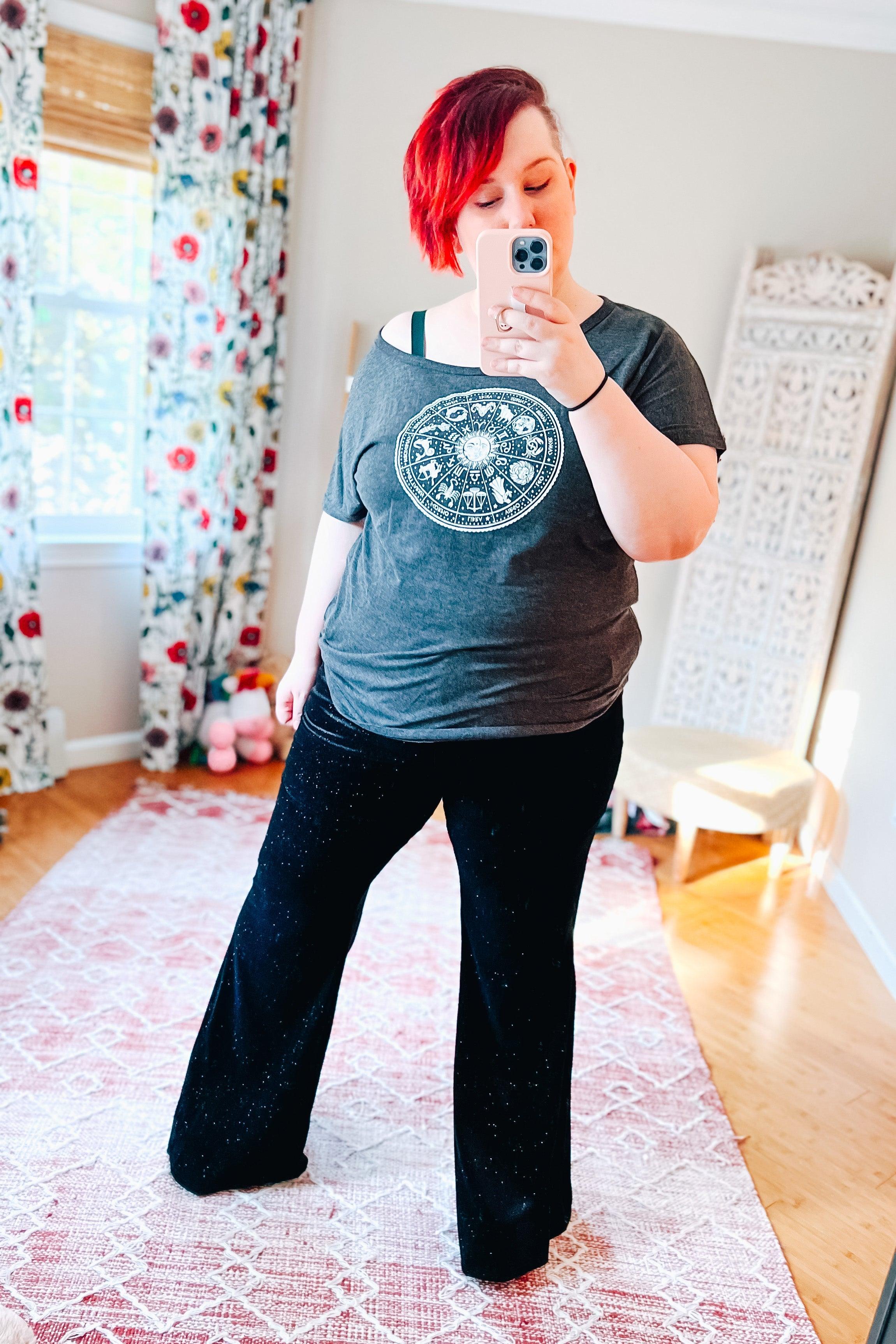 Woman with short red hair wearing black glitter flared bell bottoms and a gray graphic tee taking a mirror selfie indoors