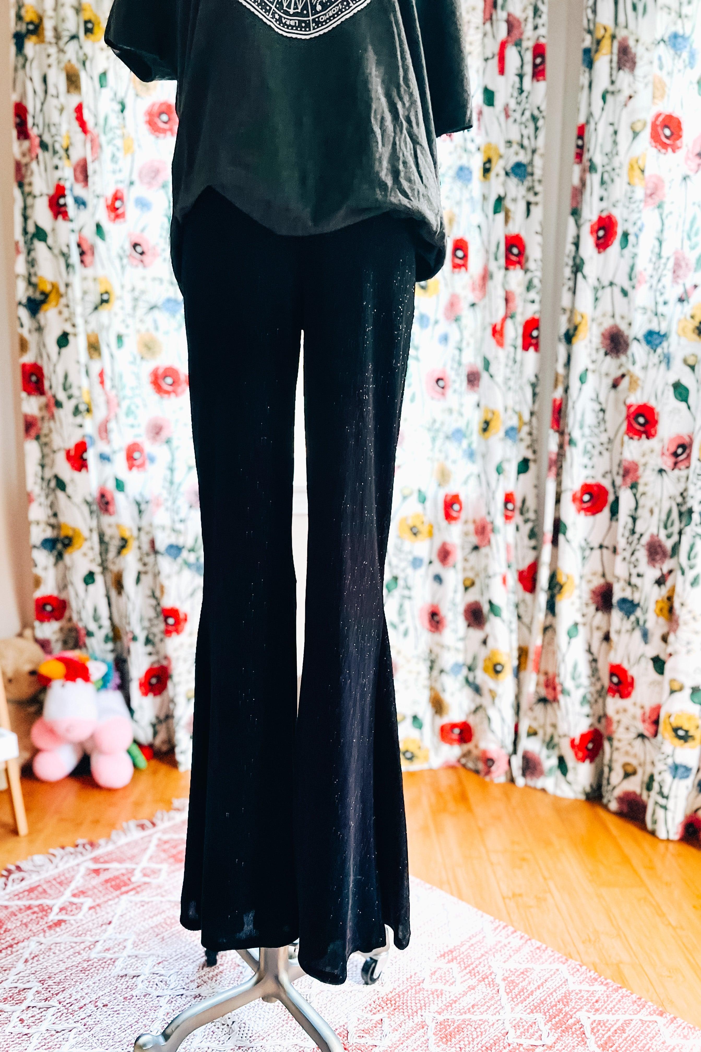 Black glitter bell bottoms displayed on a mannequin with floral curtains and wooden floor background