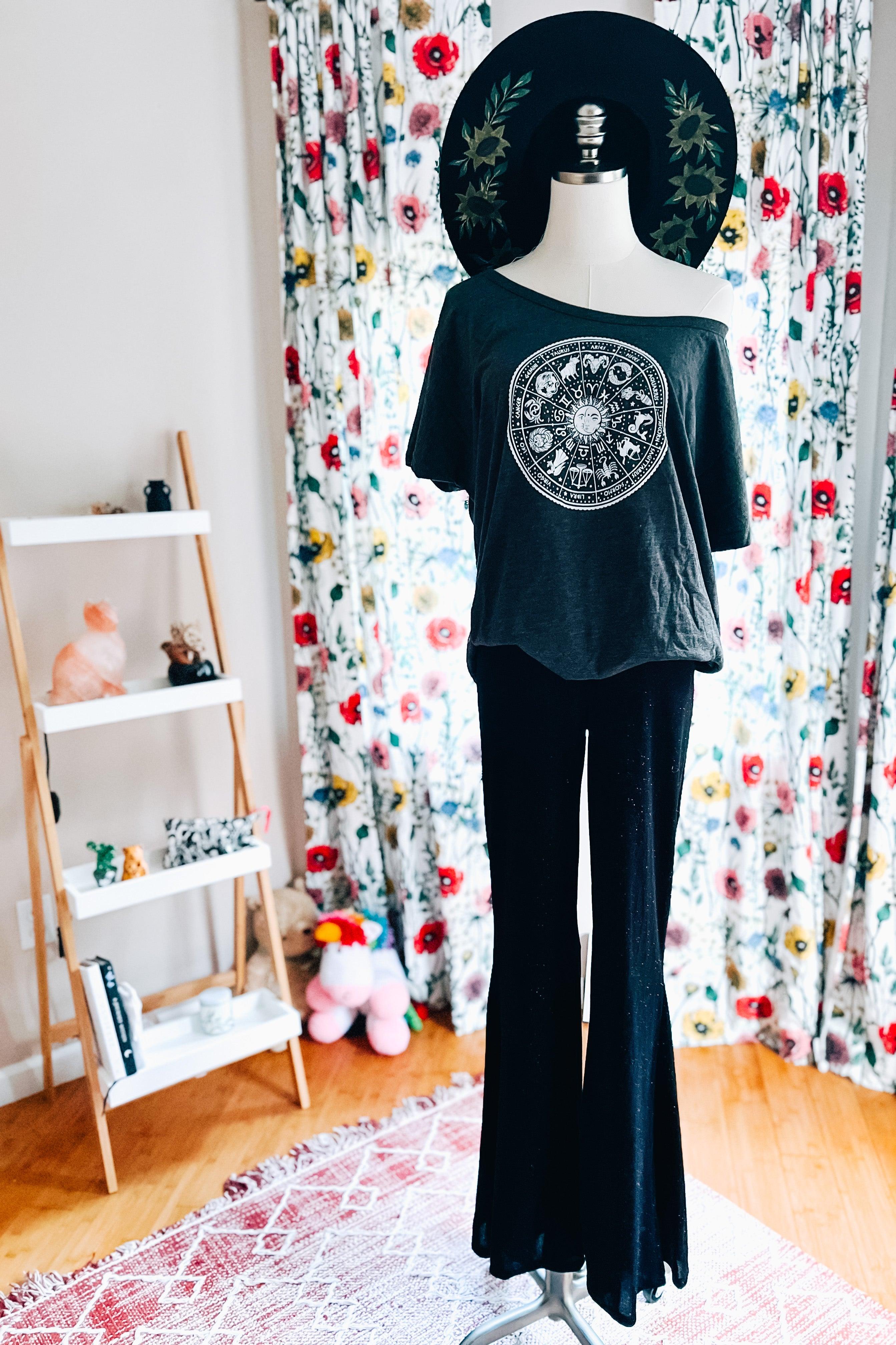 Mannequin wearing black glitter bell bottoms and loose off-shoulder black top with circular zodiac print indoors