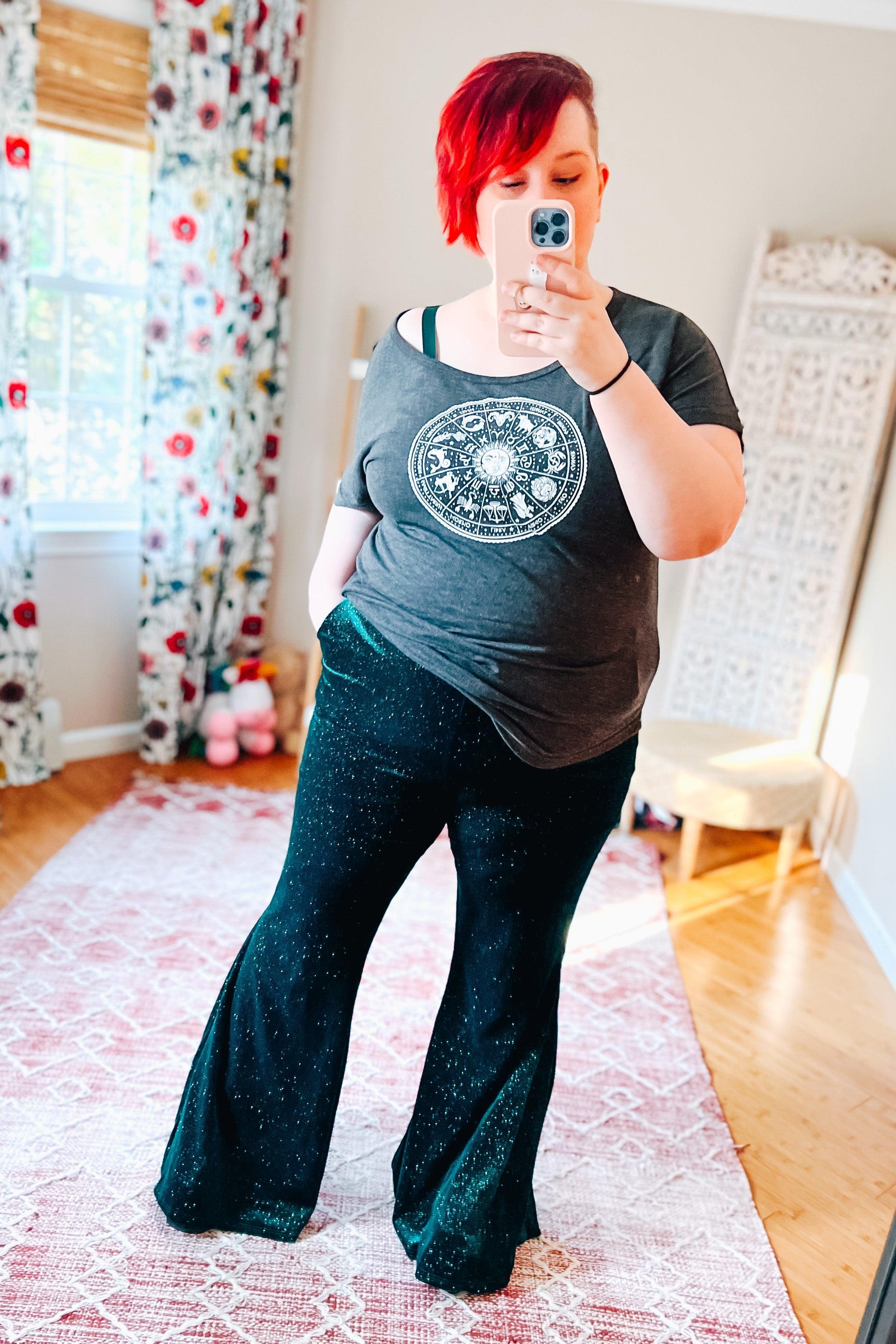 Woman taking mirror selfie wearing black glitter bell bottoms and dark grey graphic zodiac t-shirt in cozy room