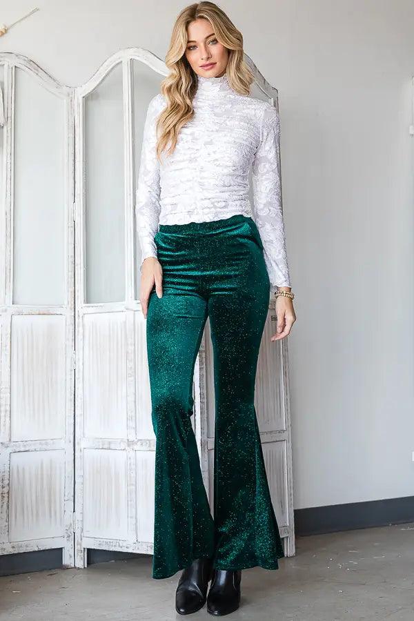 Woman modeling shimmering evergreen velvet bell bottoms with a high waist paired with a white textured long sleeve top