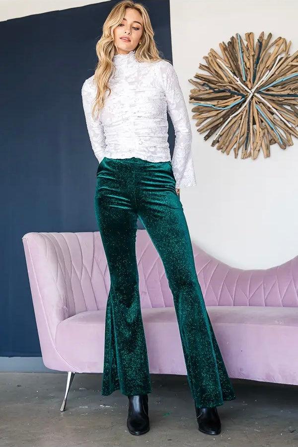 Woman wearing emerald green glitter velvet bell bottoms with a white lace long-sleeve top and black ankle boots indoors