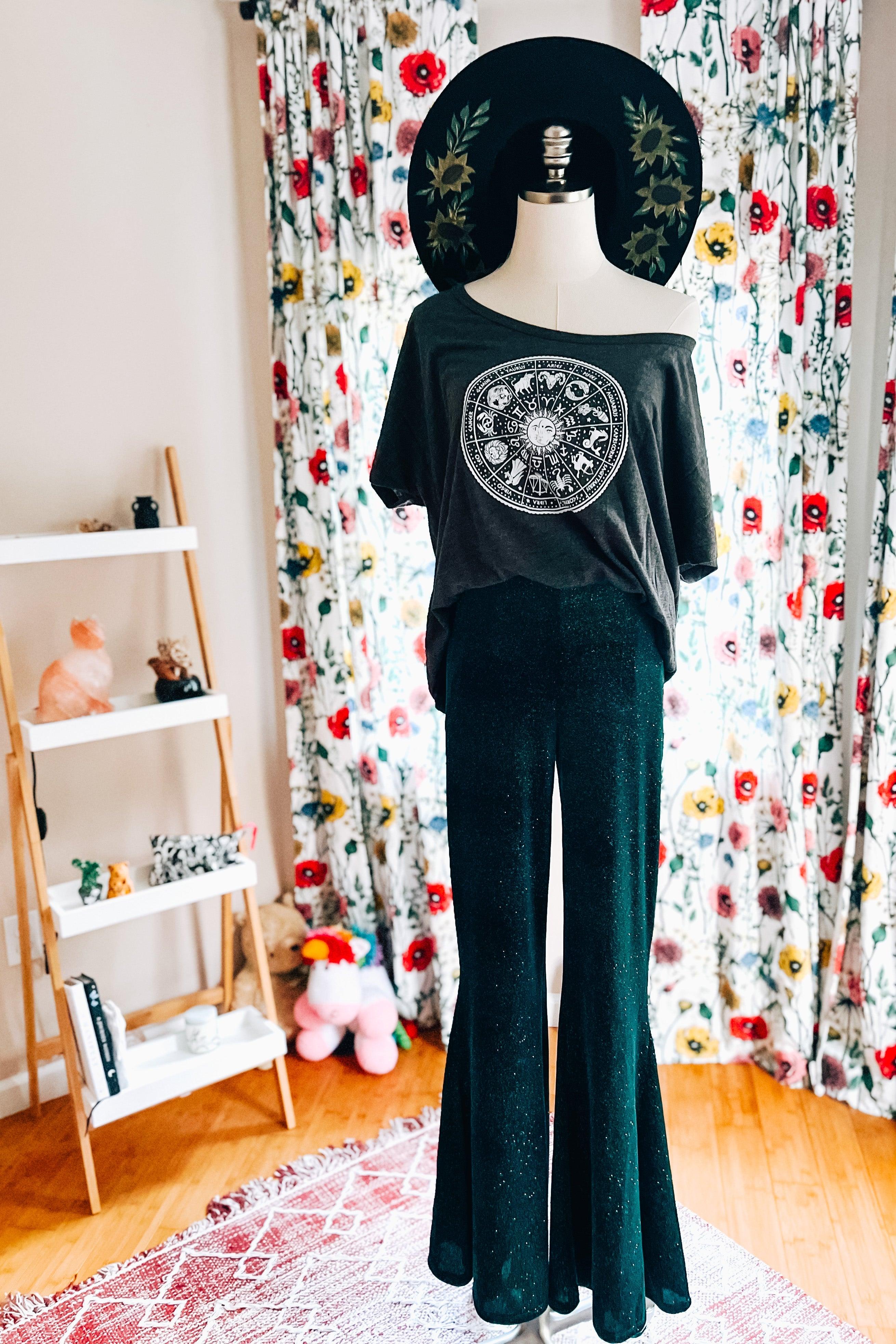 Mannequin dressed in glittery evergreen bell bottoms and loose dark grey zodiac graphic tee with floral curtains background