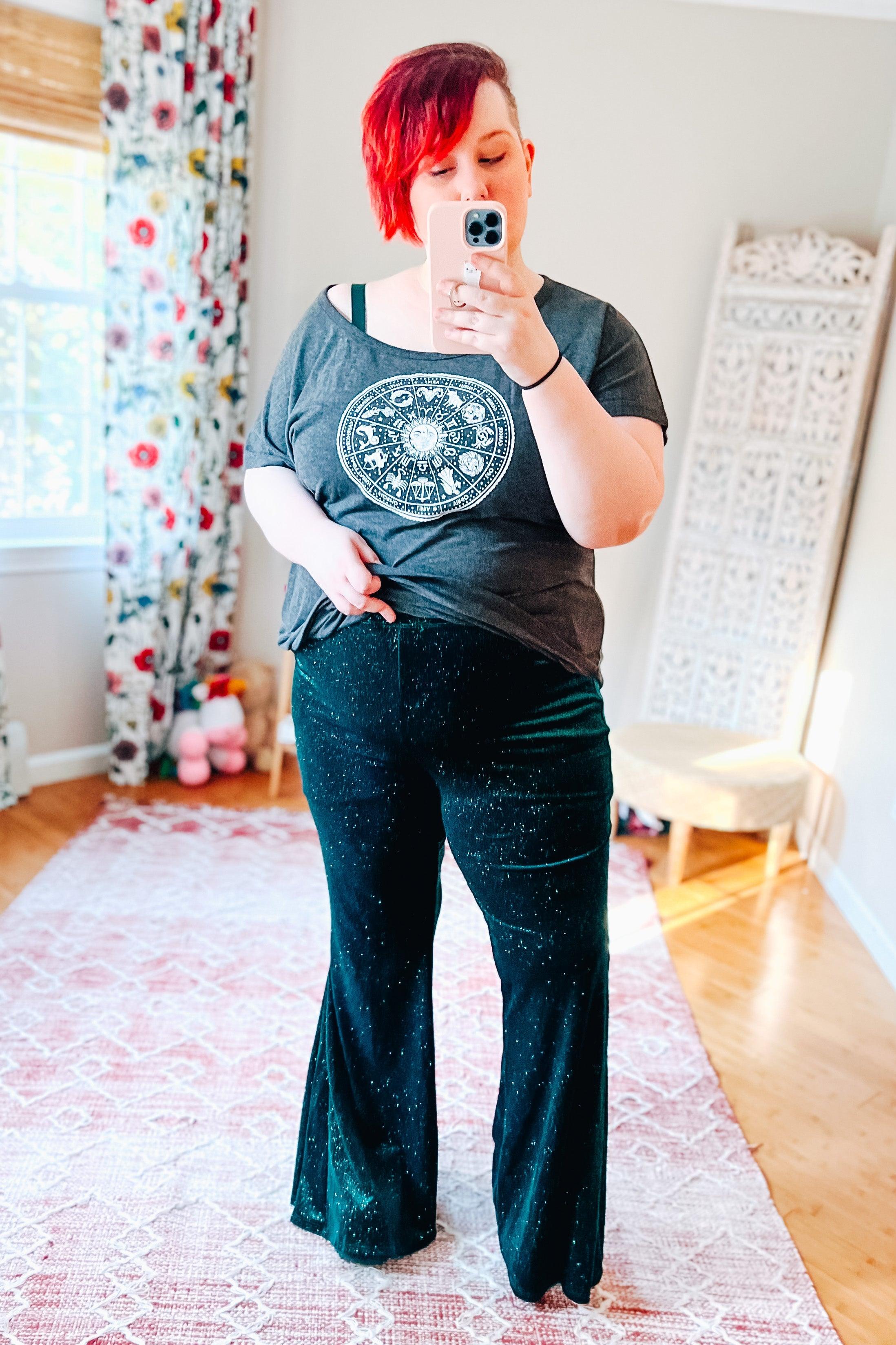 Woman wearing dark green glitter velvet bell bottoms with a loose black graphic zodiac t-shirt and visible black bra strap indoors