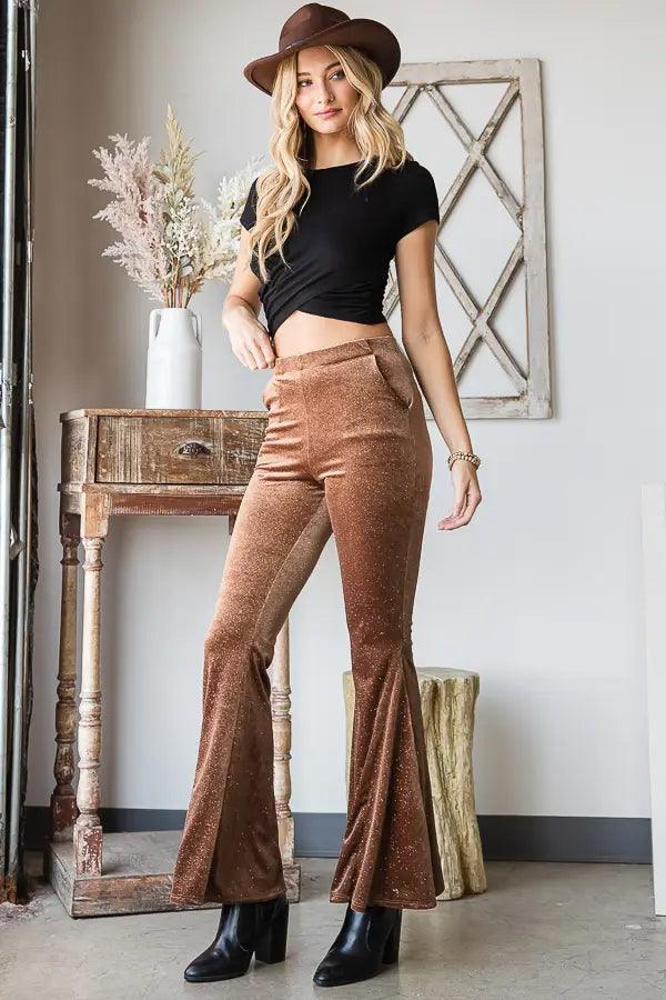 Woman standing indoors wearing bronze glitter bell bottoms with black crop top, brown hat, and black heeled boots