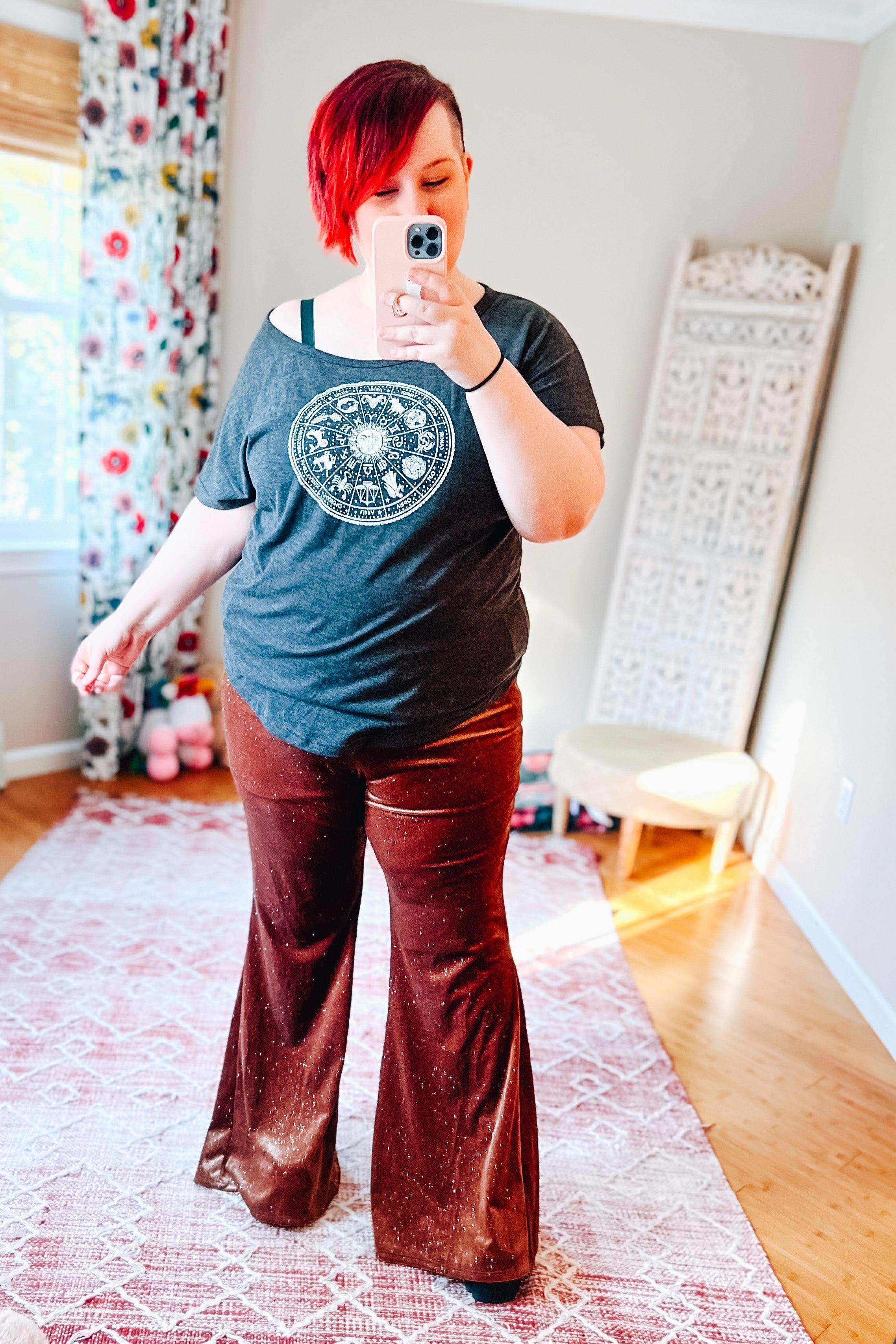 Woman wearing rust glitter velvet bell bottoms and dark zodiac graphic tee taking mirror selfie in bright room with patterned curtains