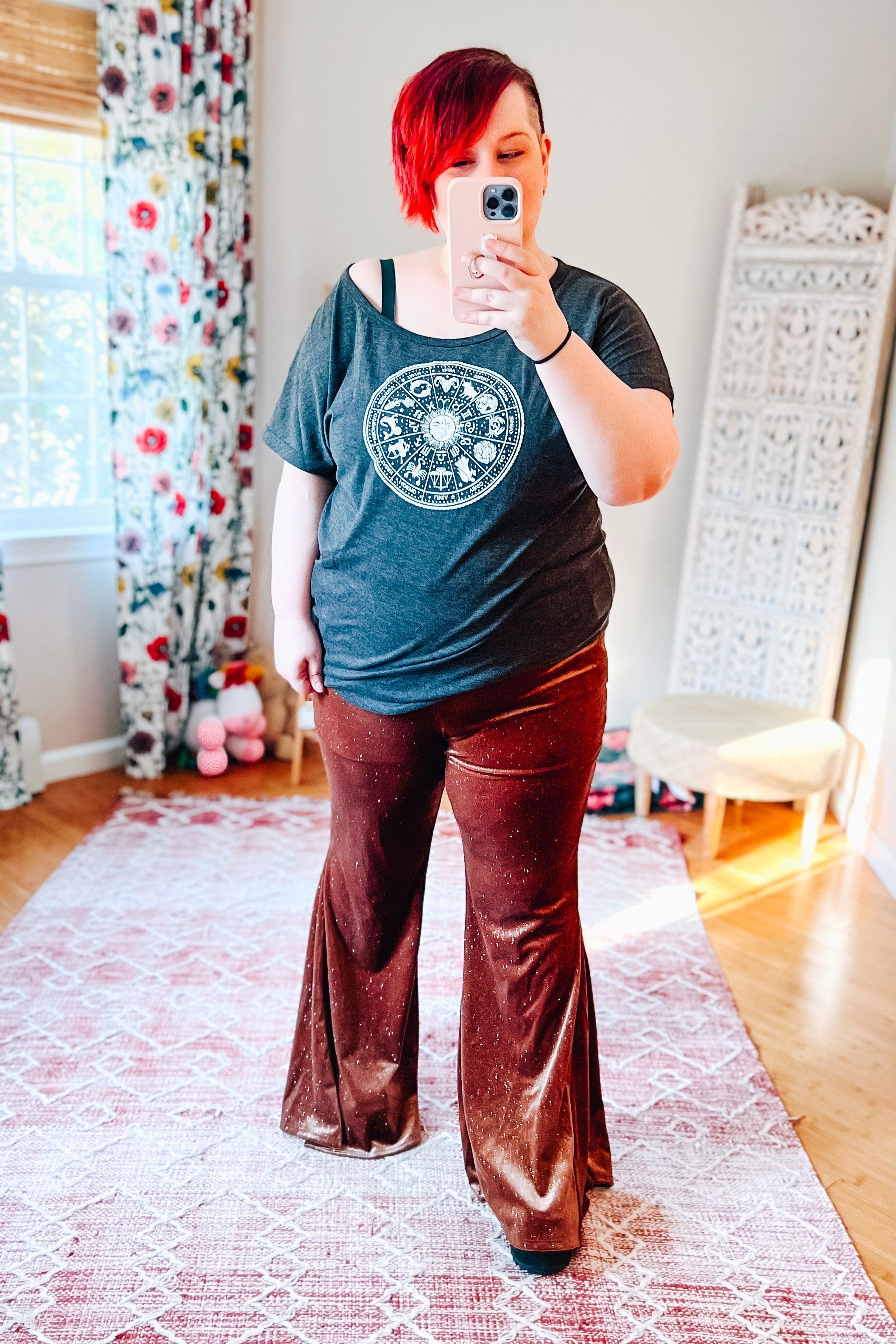 Woman taking selfie wearing rust velvet glitter bell bottoms and dark zodiac print t-shirt in bright room with floral curtains