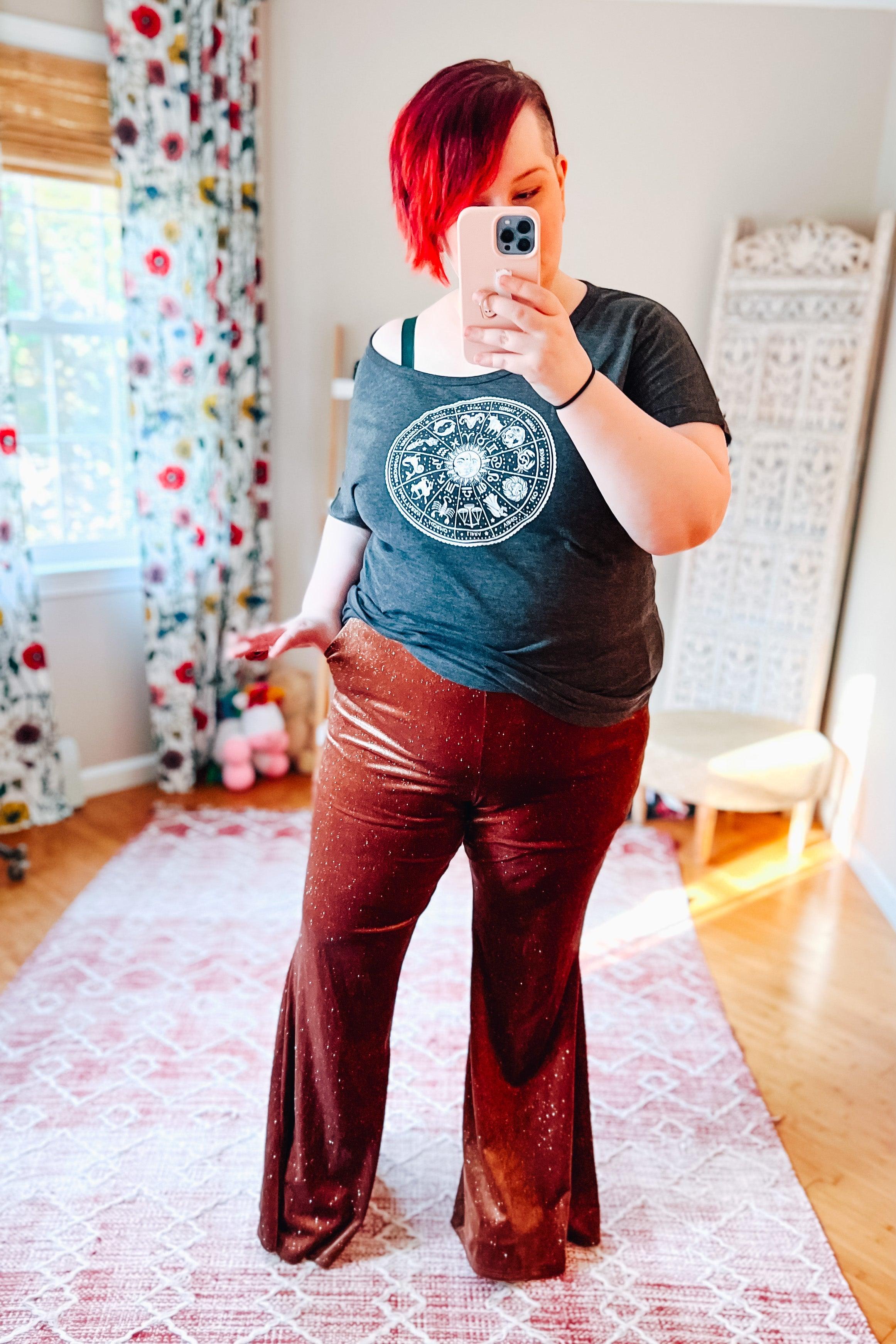 Woman taking mirror selfie wearing rust-colored glitter bell bottoms and black graphic t-shirt in sunlit room with floral curtains