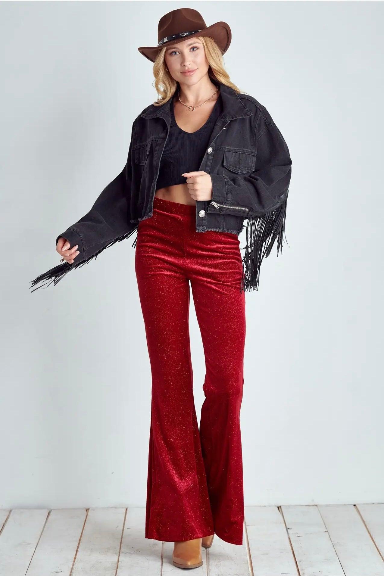 Woman wearing ruby red glitter bell bottoms paired with cropped black fringe jacket and brown hat on white floor background