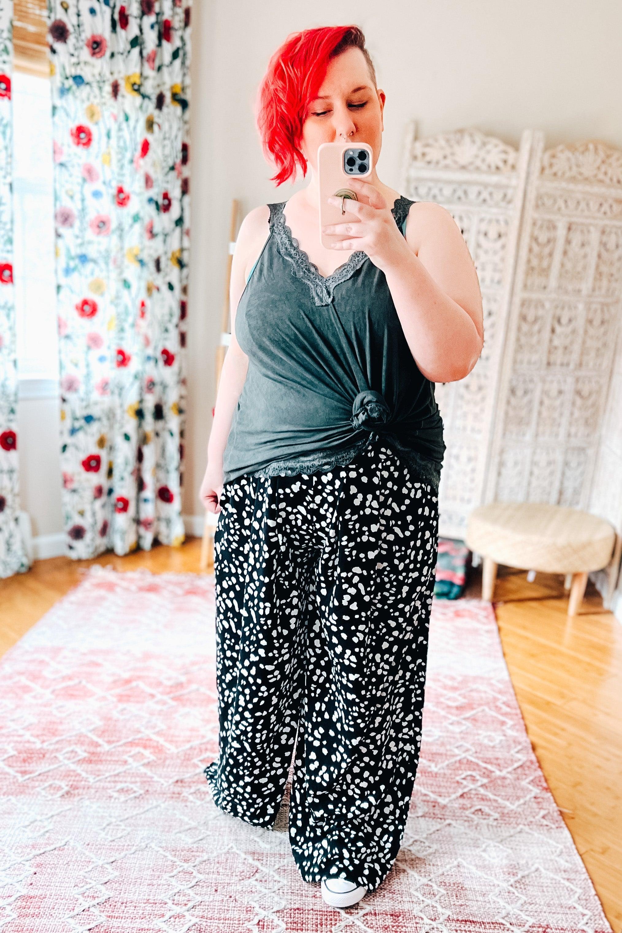 Woman with red hair wearing black wide leg pants with white Dalmatian spots and a knotted dark gray tank top taking selfie indoors