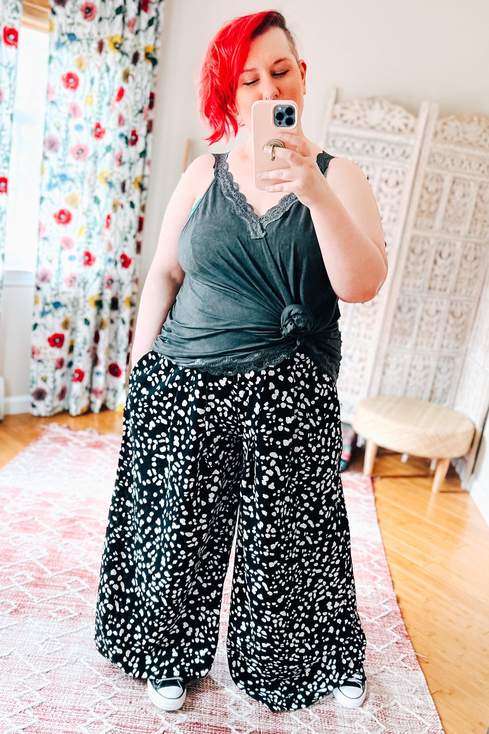 Woman with bright red hair wearing black wide leg pants with white abstract spots and dark gray tank top taking mirror selfie