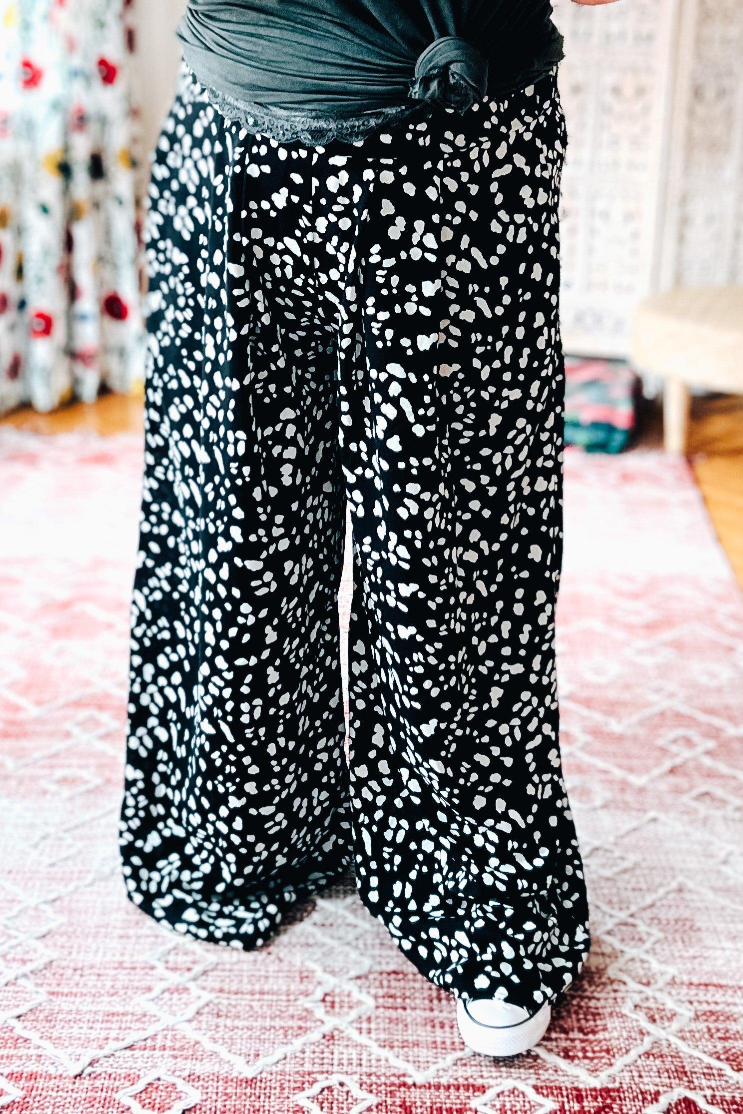 Close-up of black wide leg pants with scattered white irregular spots worn with a tied black top and white sneakers