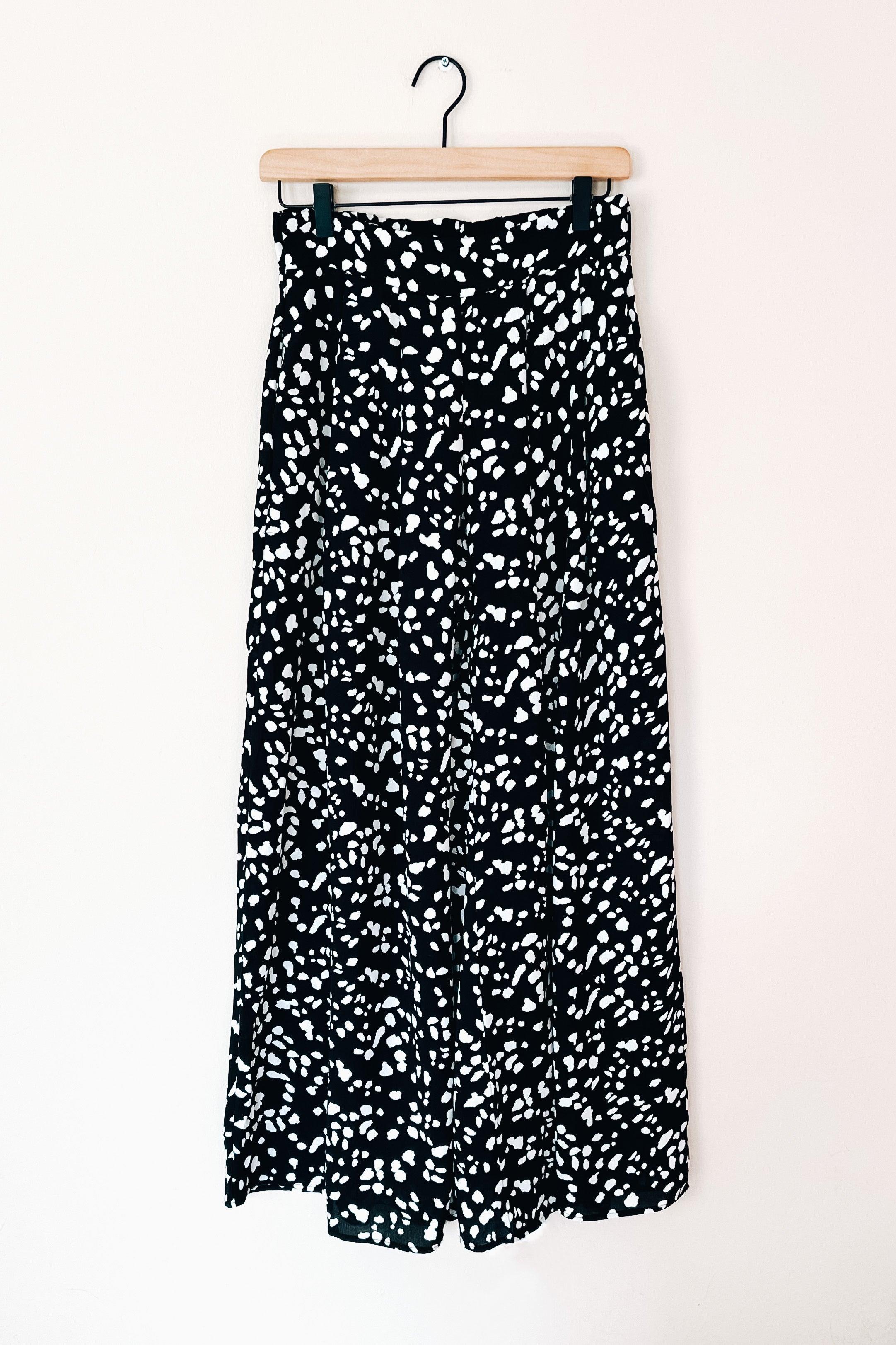 Black wide leg pants featuring a white irregular dot pattern hanging on wooden hanger against white wall