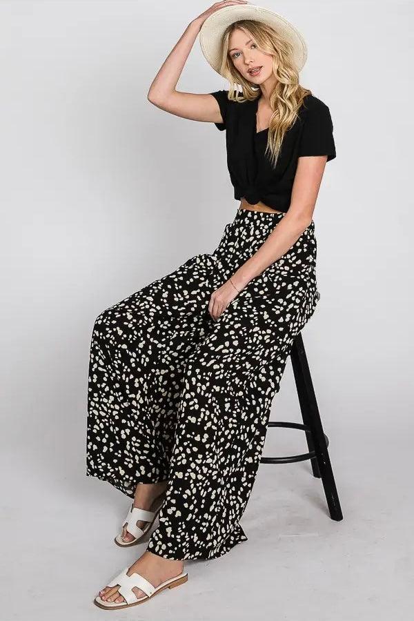 Woman seated on stool wearing black wide leg pants with scattered white spot pattern, black knotted top, straw hat, and white sandals