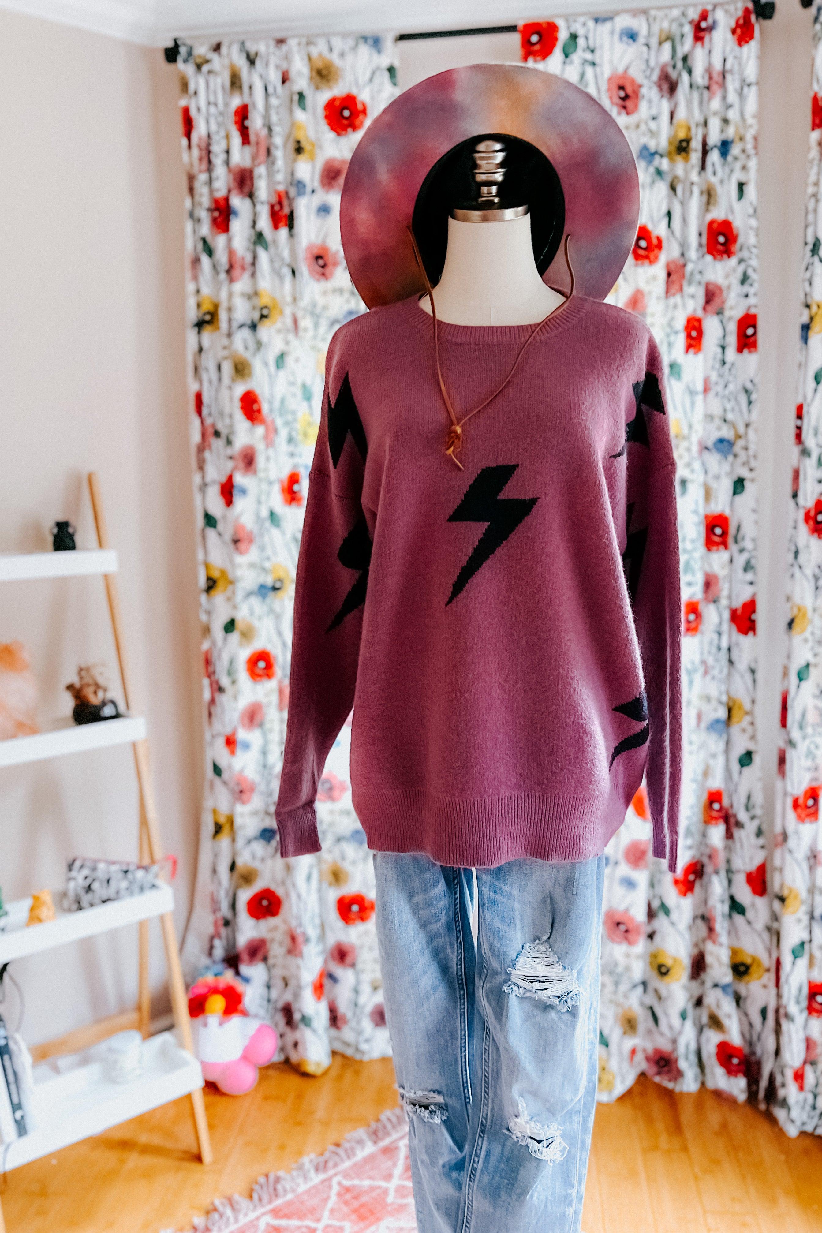 Dress form wearing a purple sweater with black lightning bolts, blue distressed jeans and a hand painted hat in an indoor setting with floral curtains
