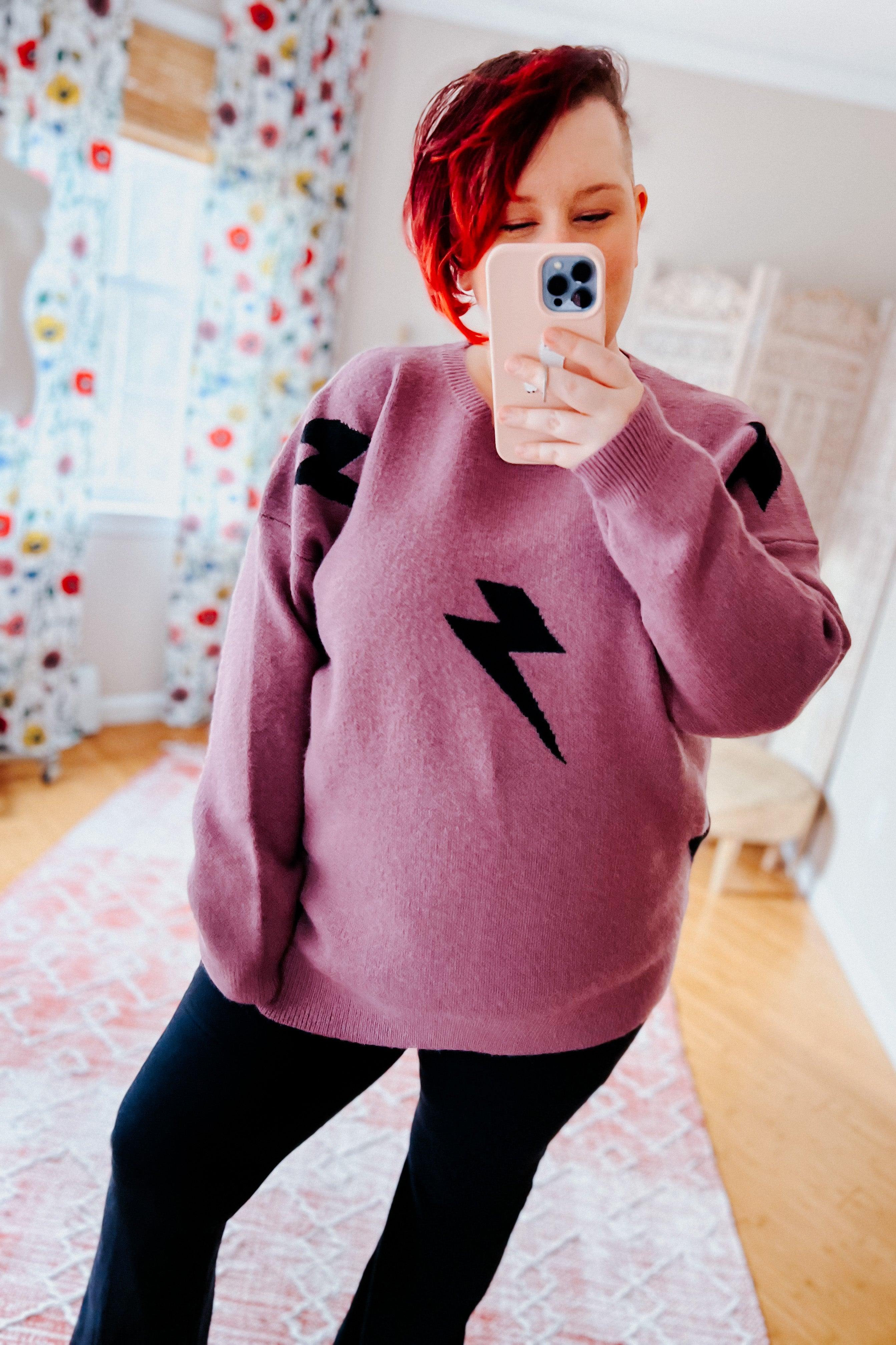 Woman wearing a purple sweater with black lightning bolt designs and black flared leggings, standing in a room with floral curtains.