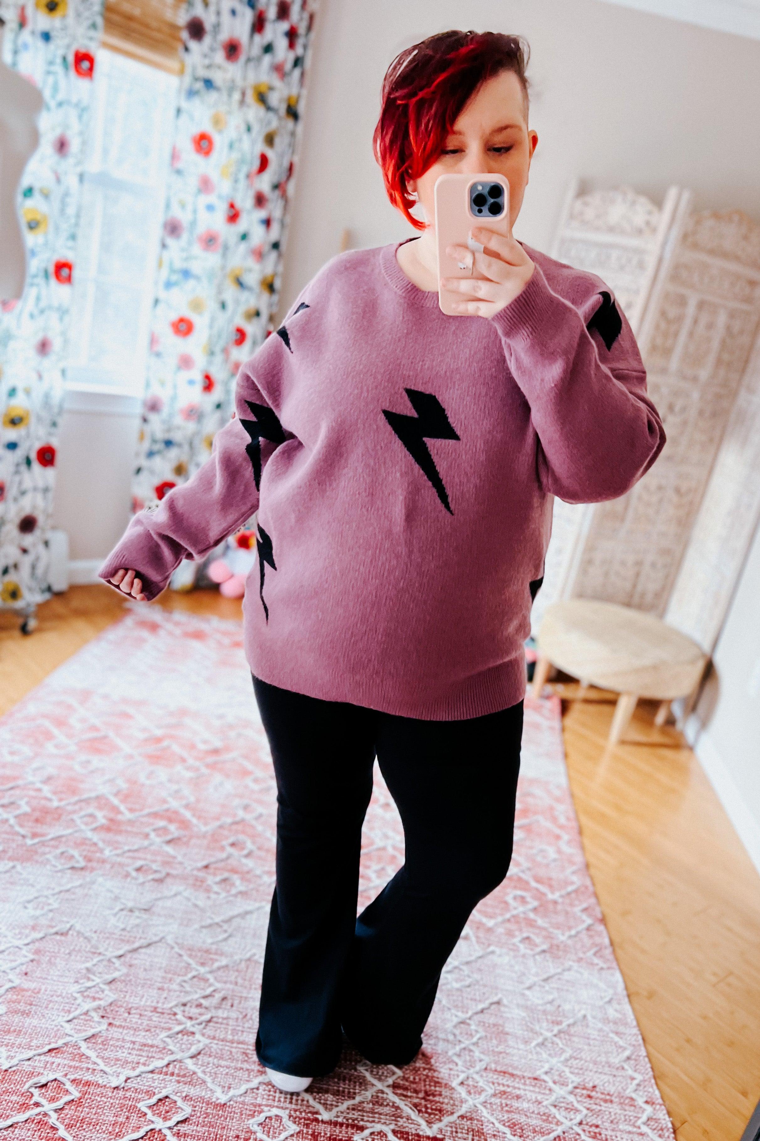 Woman wearing a purple sweater with black lightning bolt designs and black flared leggings, standing in a room with floral curtains.