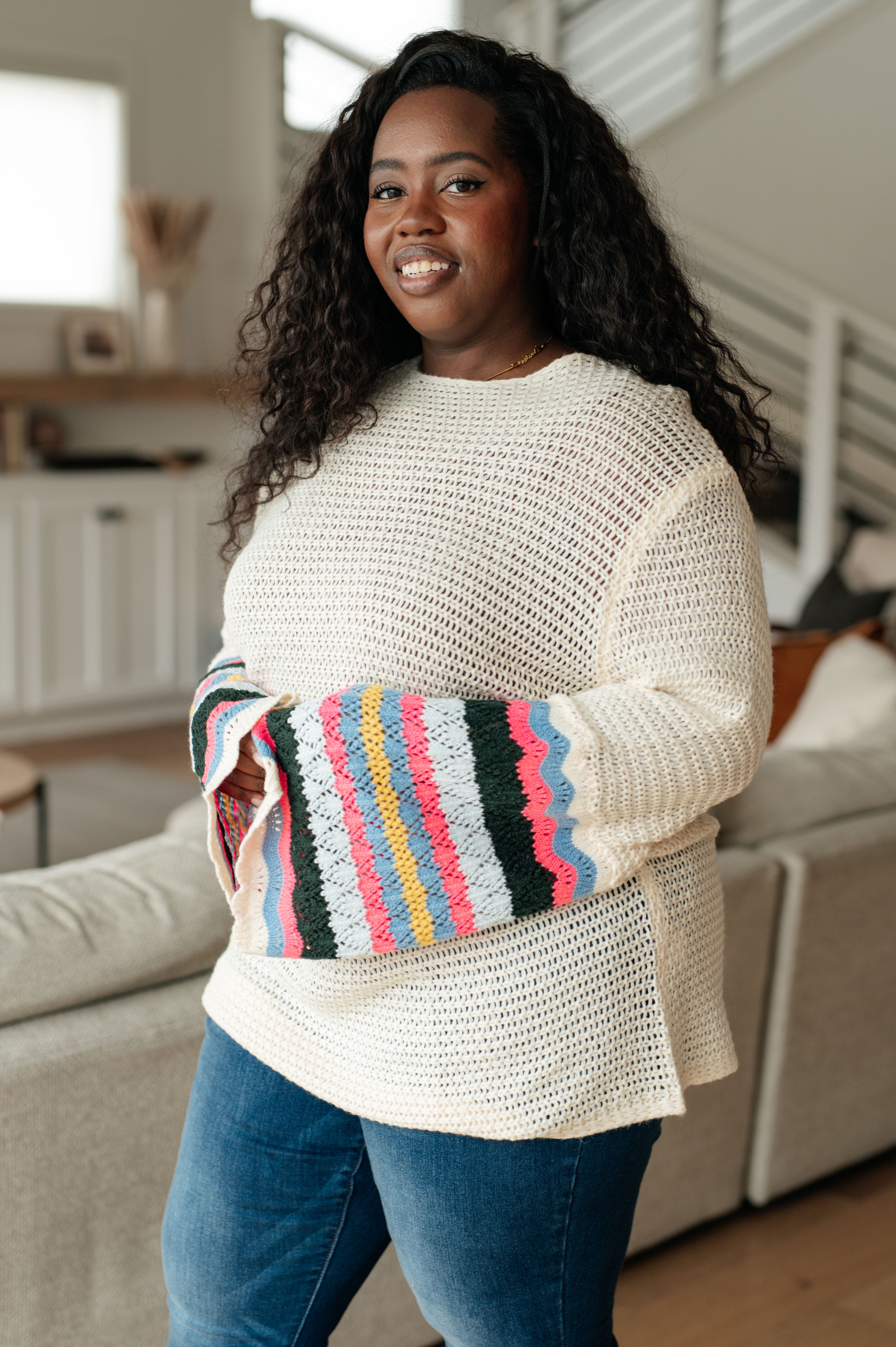 Smiling woman indoors wearing cream knit sweater with colorful striped wide sleeves and blue jeans standing by couch