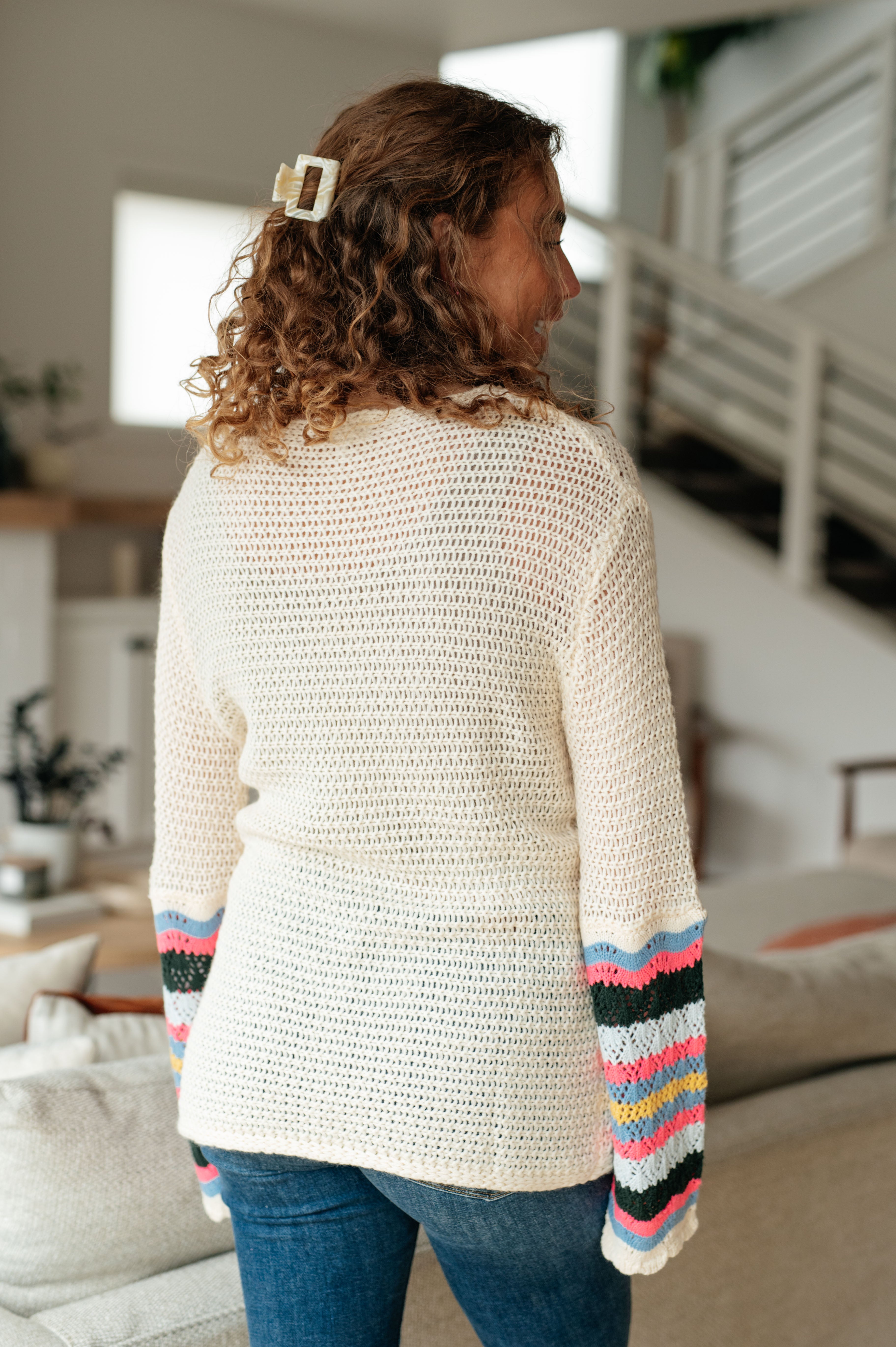 Woman wearing cream crochet sweater with colorful striped sleeves and blue jeans viewed from back indoors