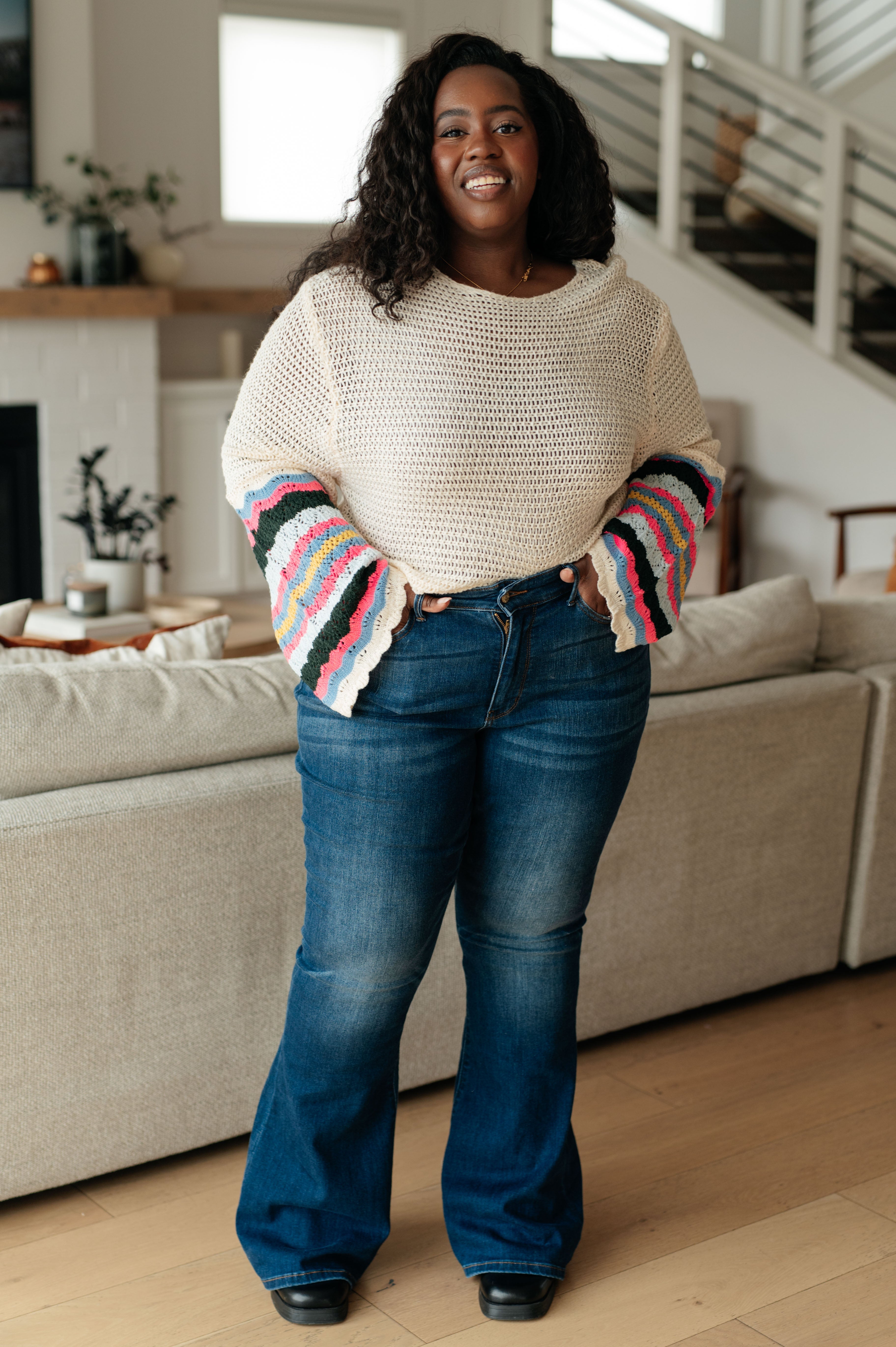 Smiling woman wearing cream sweater with colorful scalloped striped sleeves and high-waisted flared jeans indoors by sofa