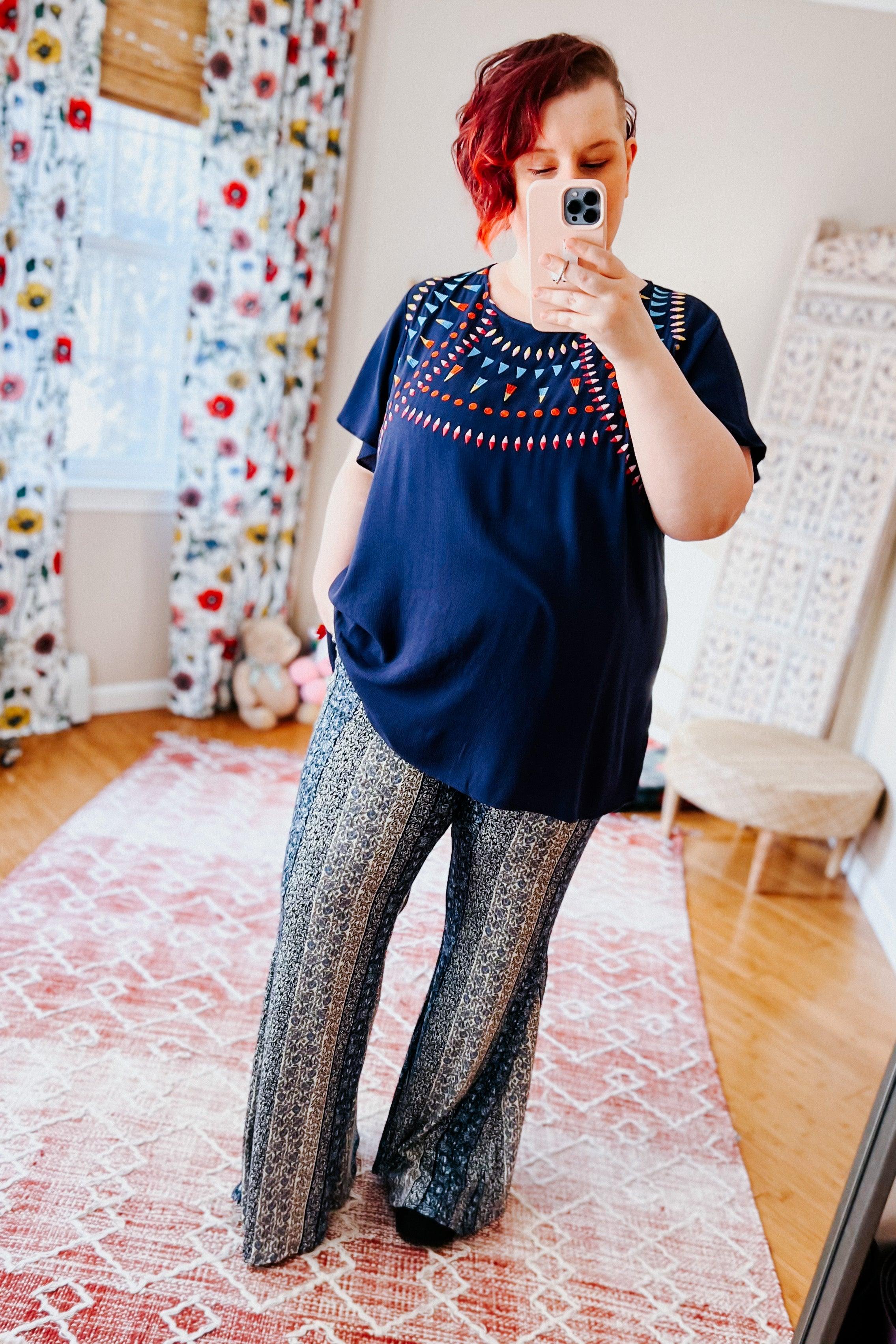 Fleetwood Floral Bell Bottoms in navy with intricate vertical patchwork patterns, styled with a loose navy top in a bright room featuring floral curtains and a soft rug.
