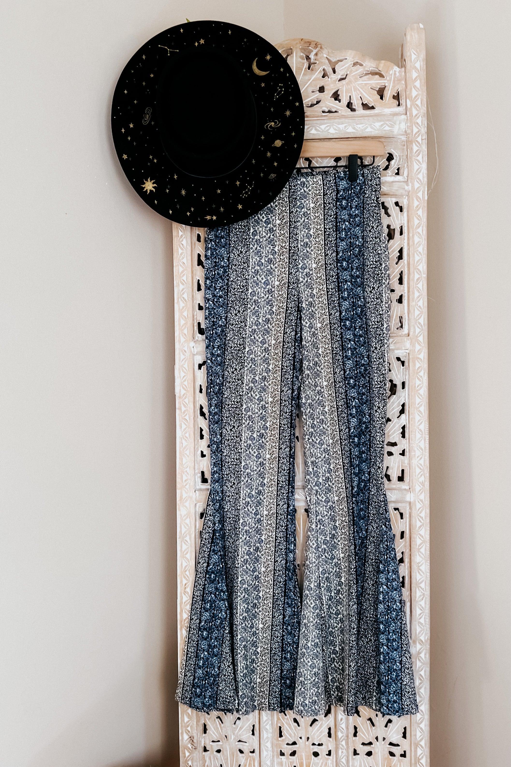Fleetwood Floral Bell Bottoms in navy with vertical patchwork floral stripes, styled hanging on a carved wood screen alongside a black hat with gold celestial accents