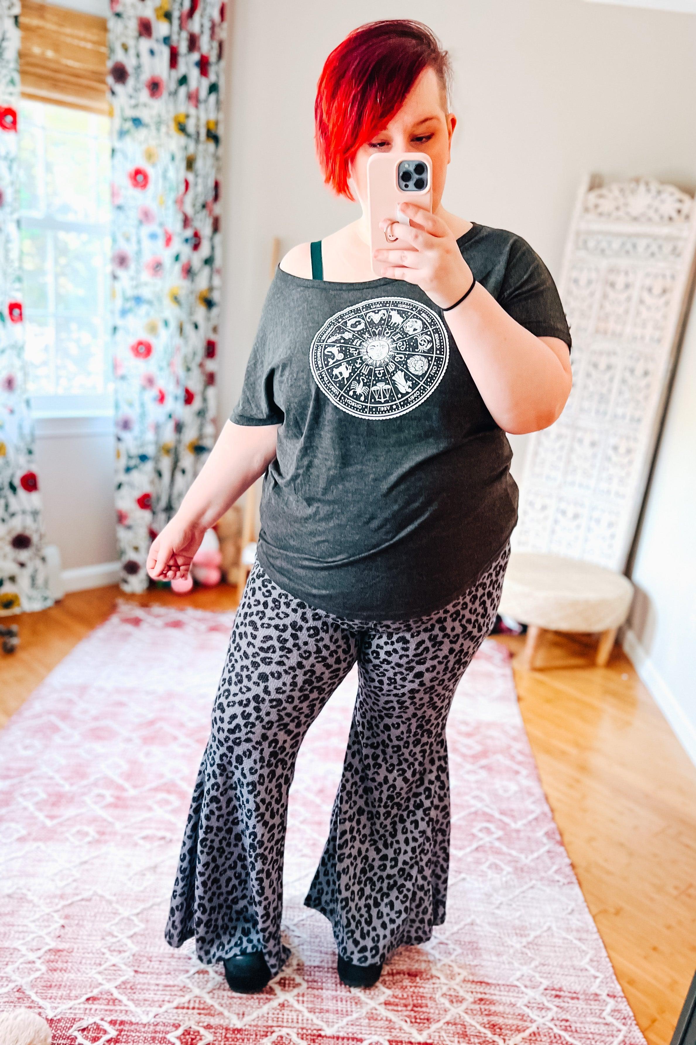 Woman taking selfie wearing black and blue leopard print bell bottom pants and dark graphic off-shoulder top indoors