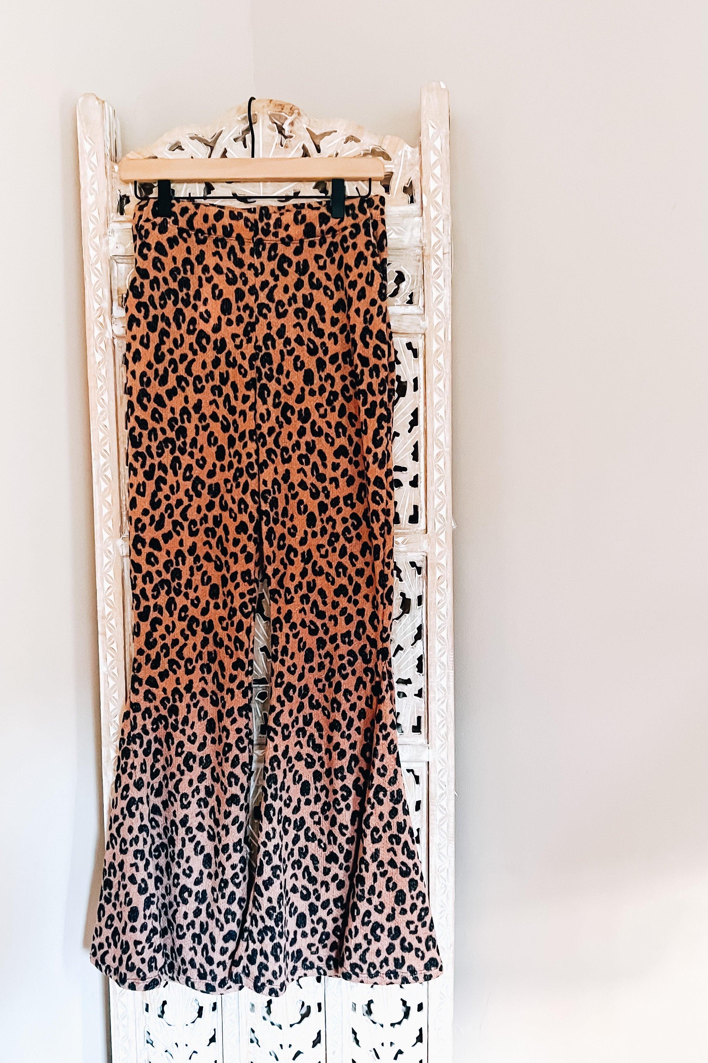 Mocha leopard print bell bottoms with flared legs hanging on wooden hanger against white carved screen background