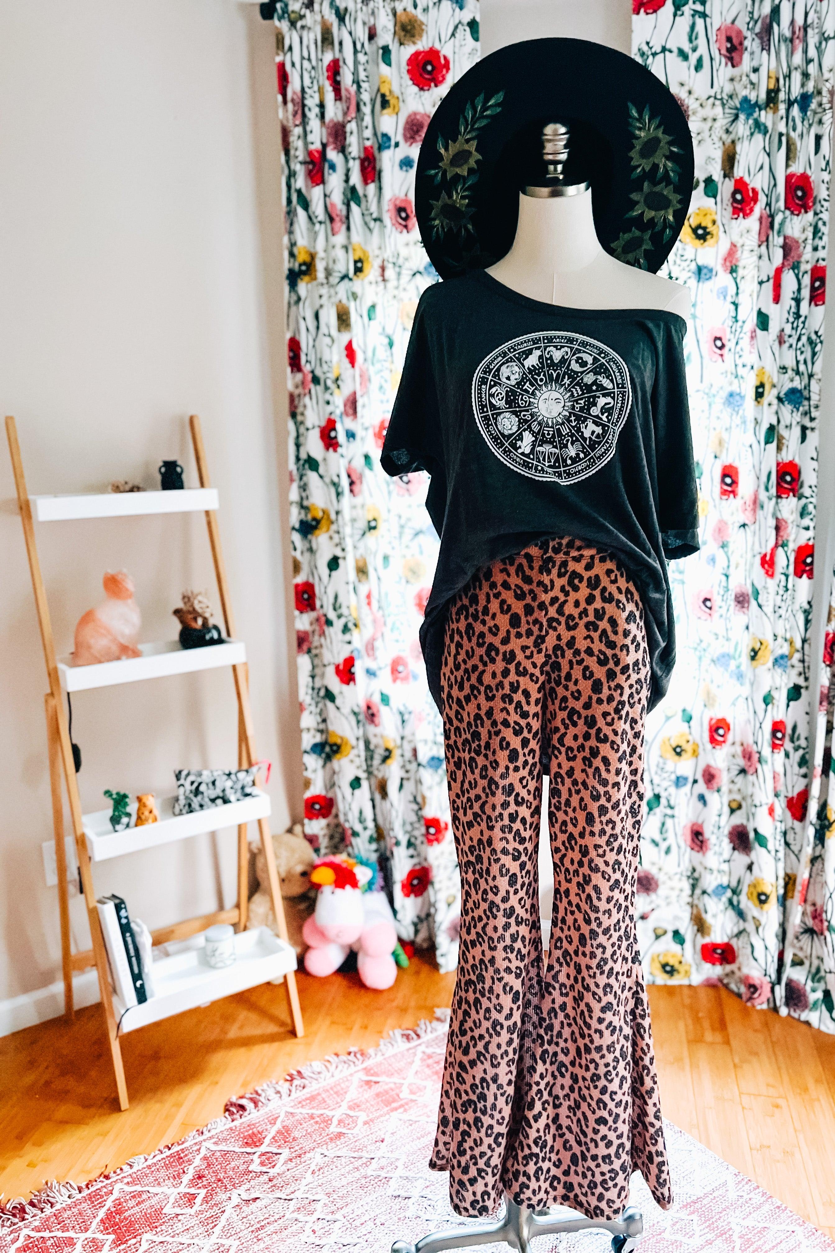 Mannequin wearing mocha animal print bell bottoms paired with black graphic off-shoulder top and wide brim hat indoors