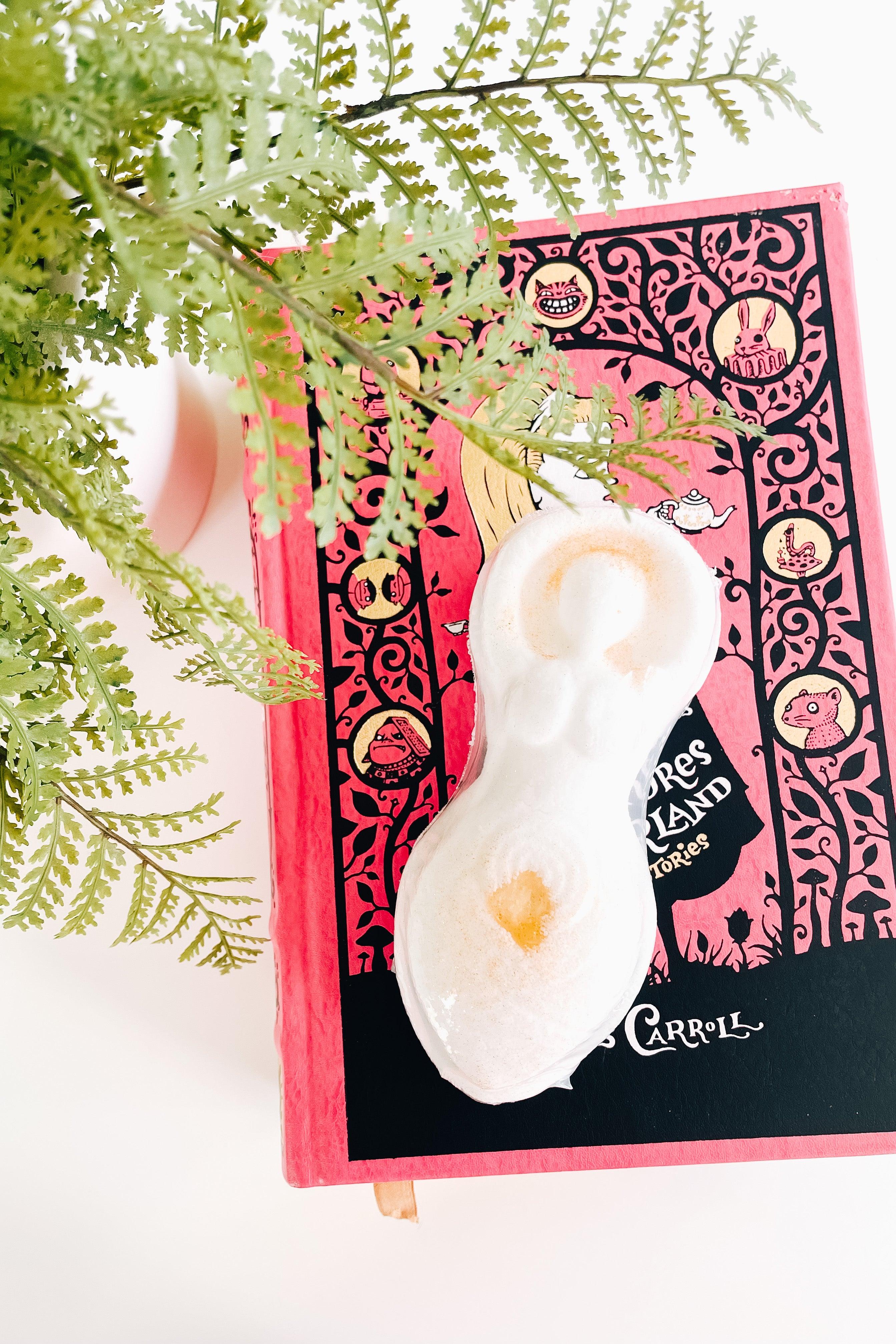 Oval-shaped cream bath bomb with golden accents resting on a pink and black illustrated book next to green fern leaves