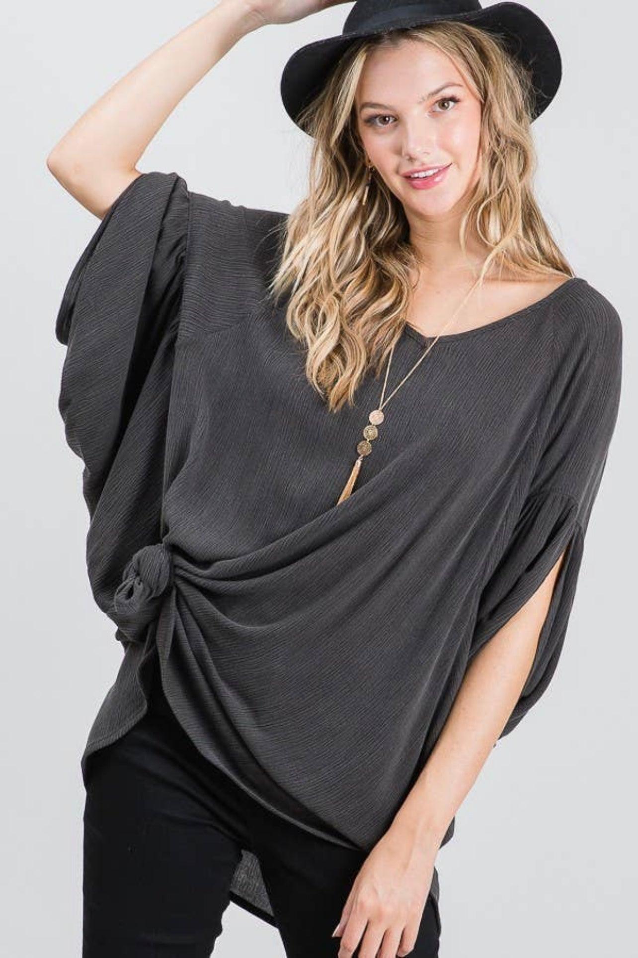 Woman wearing charcoal flowy tunic with knotted side detail and wide sleeves paired with black hat and pants