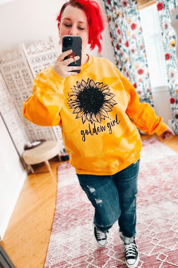 Woman with red hair taking selfie wearing yellow bleached pullover with large black sunflower and golden girl text indoors