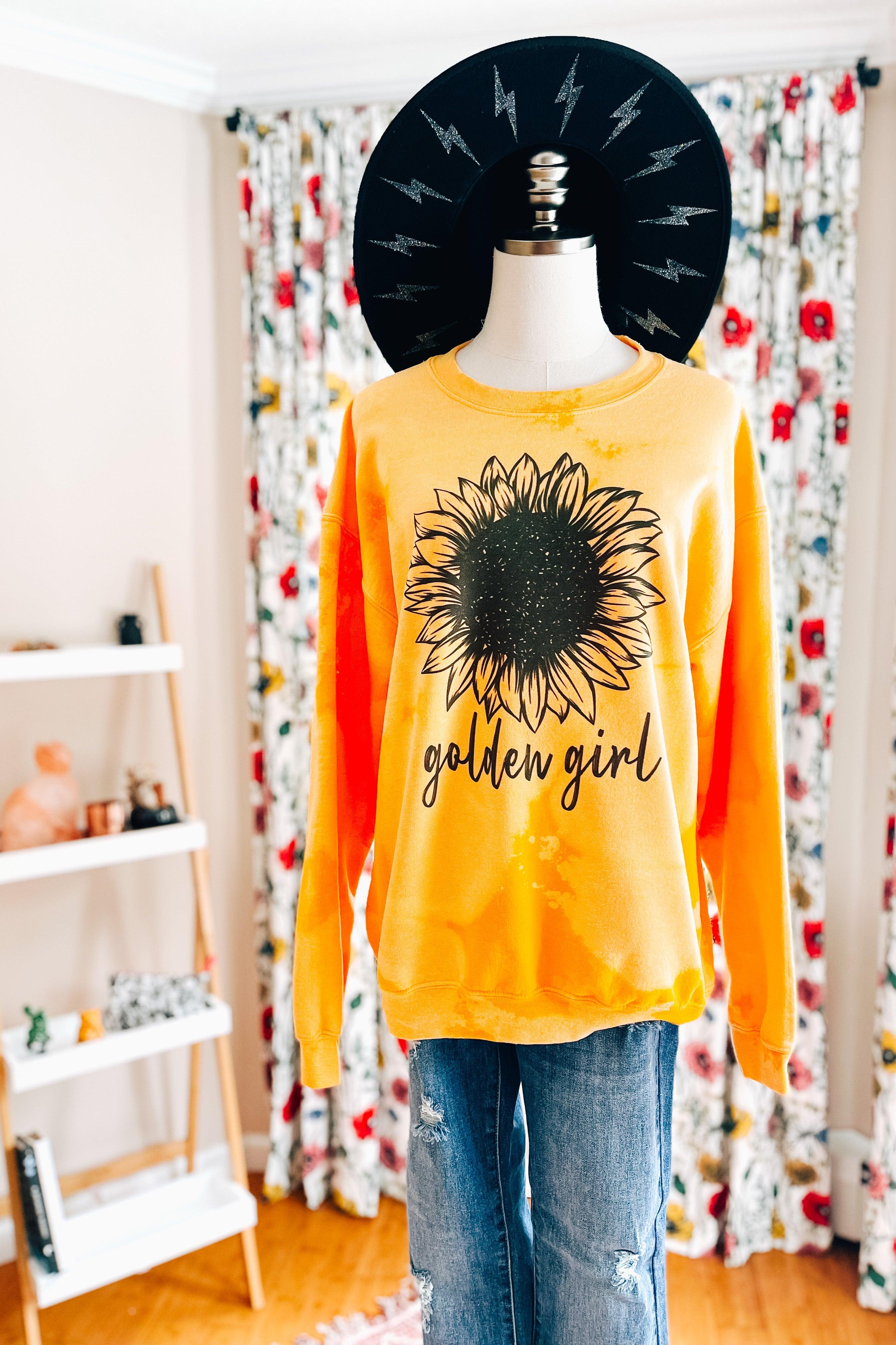 Yellow bleached pullover featuring a large black sunflower and golden girl text displayed on a mannequin with jeans indoors