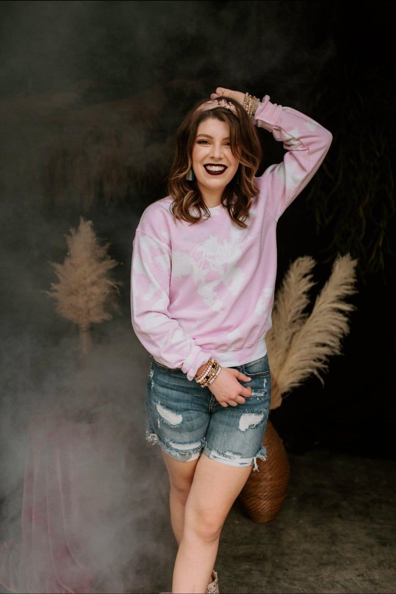 Smiling woman wearing pink tie-dye pullover with white script paired with distressed denim shorts posing indoors