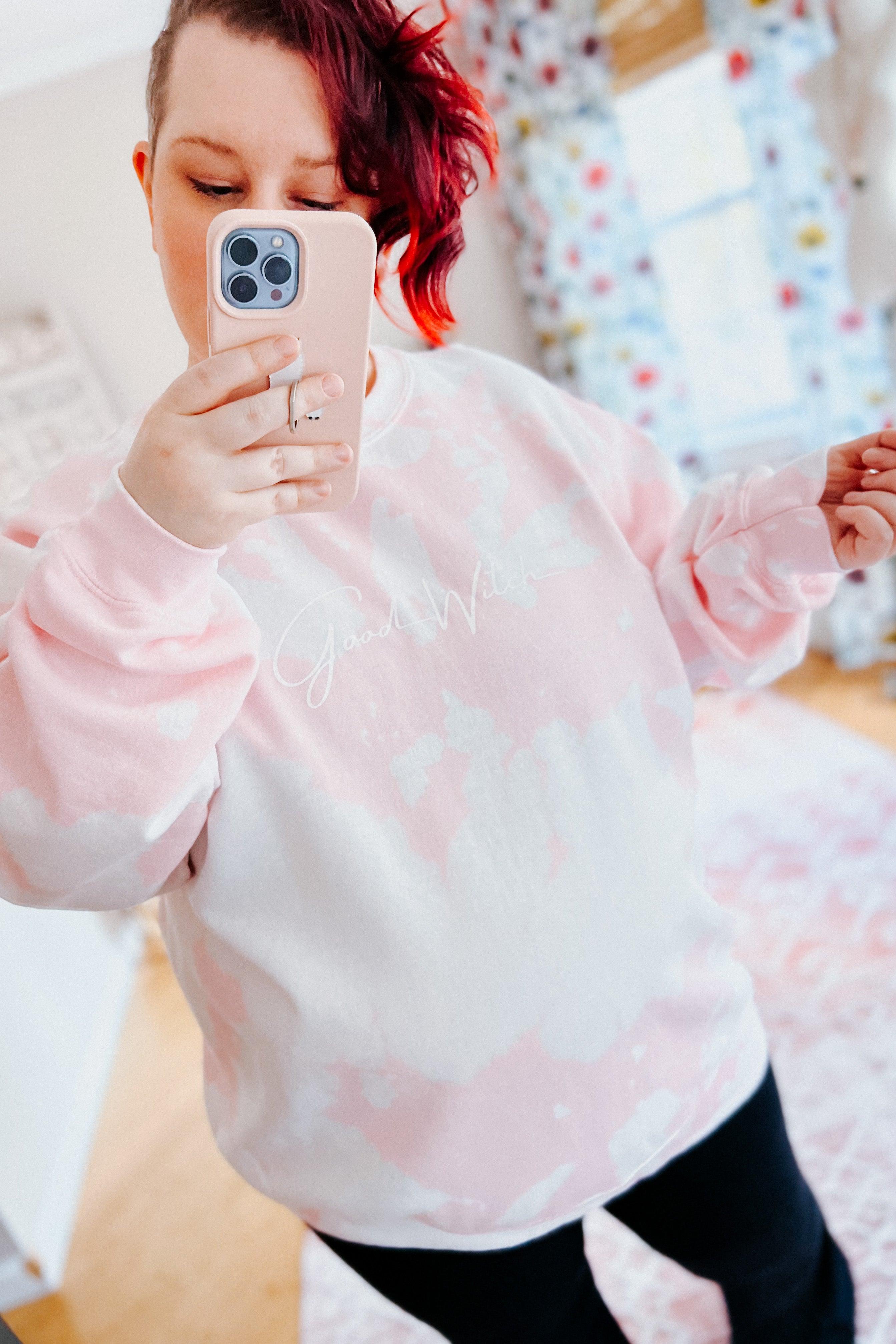 Woman taking selfie wearing pink and white bleached pullover with white Good Witch script in cozy room