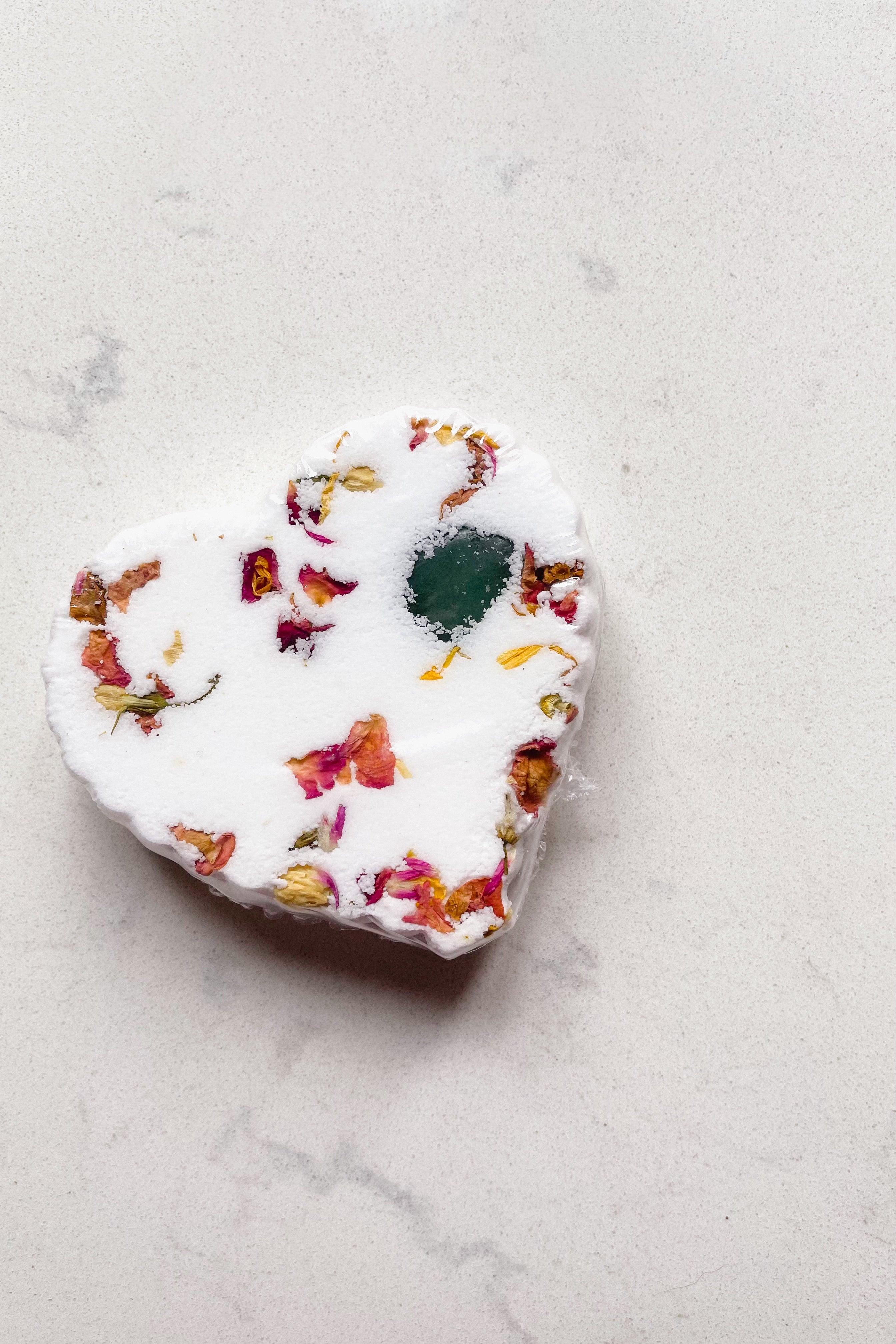 White heart-shaped bath bomb with dried flower petals and green crystal detail on light marble surface