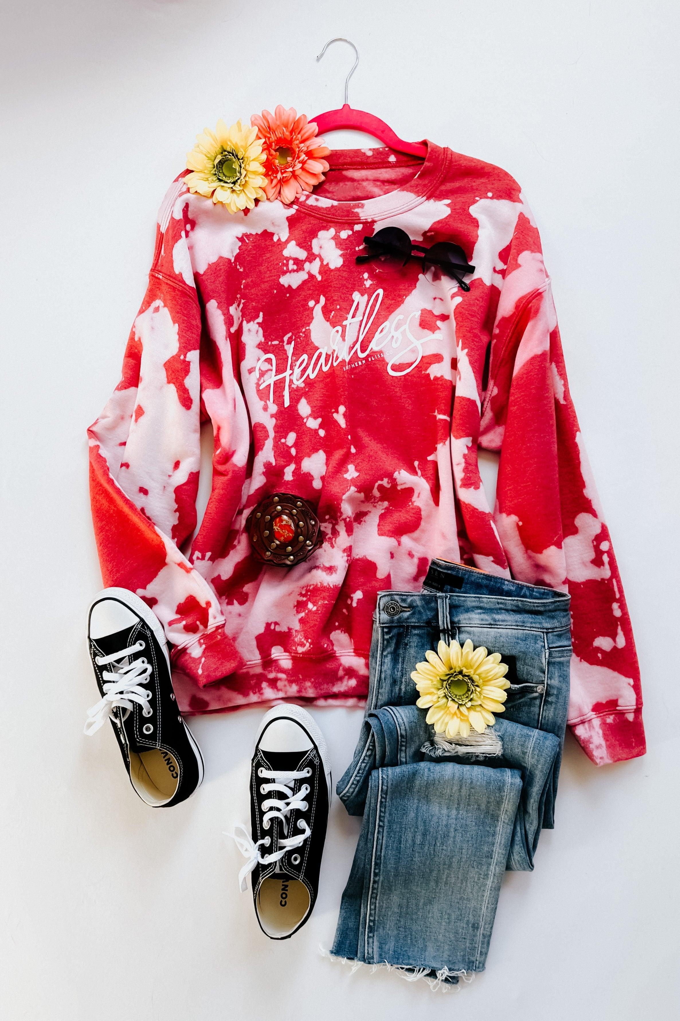 Red bleached pullover with white "Heartless" script logo styled with denim jeans, black sneakers, and floral accents on white background