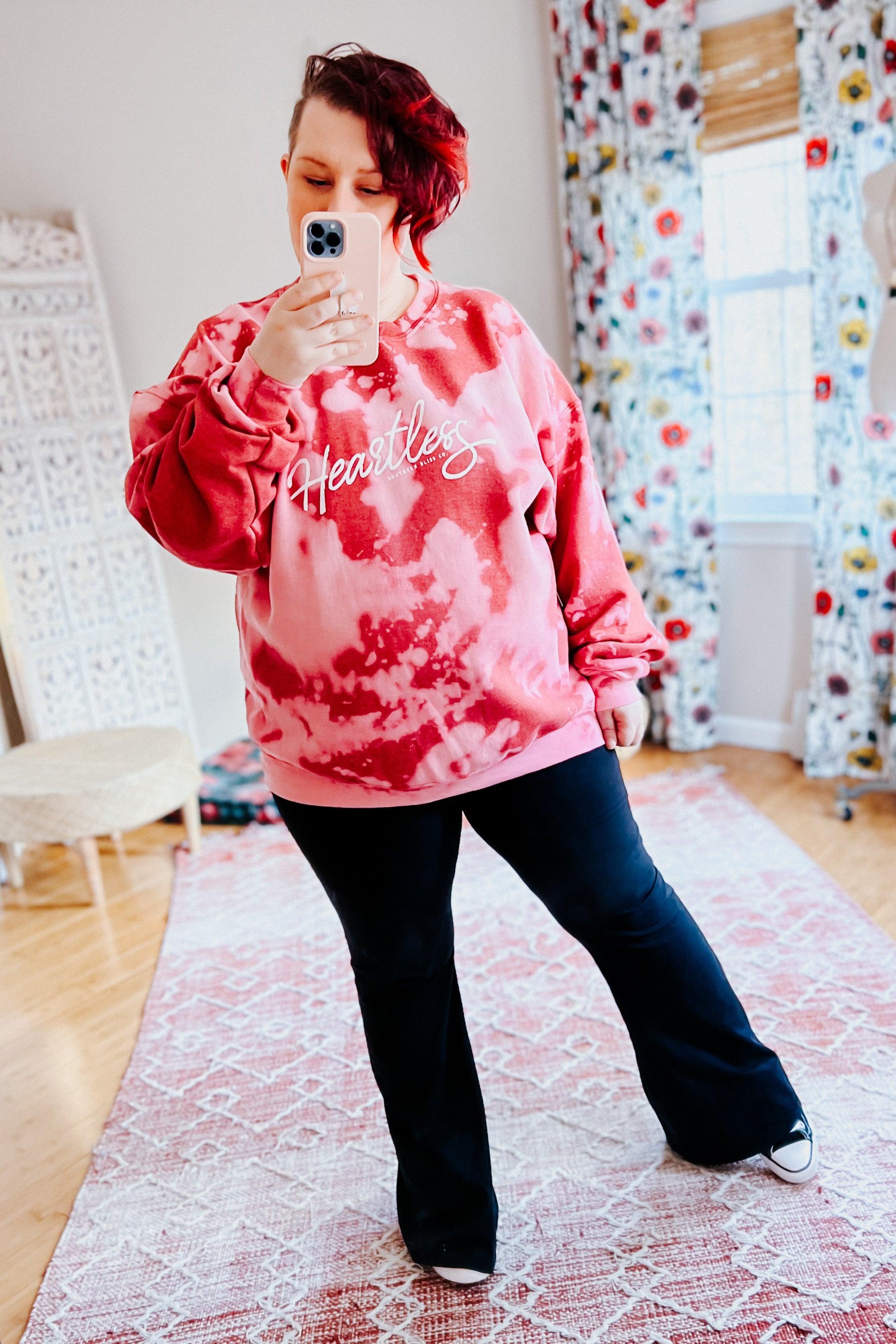 Woman taking mirror selfie wearing red heartless bleached pullover with white script logo and black pants indoors