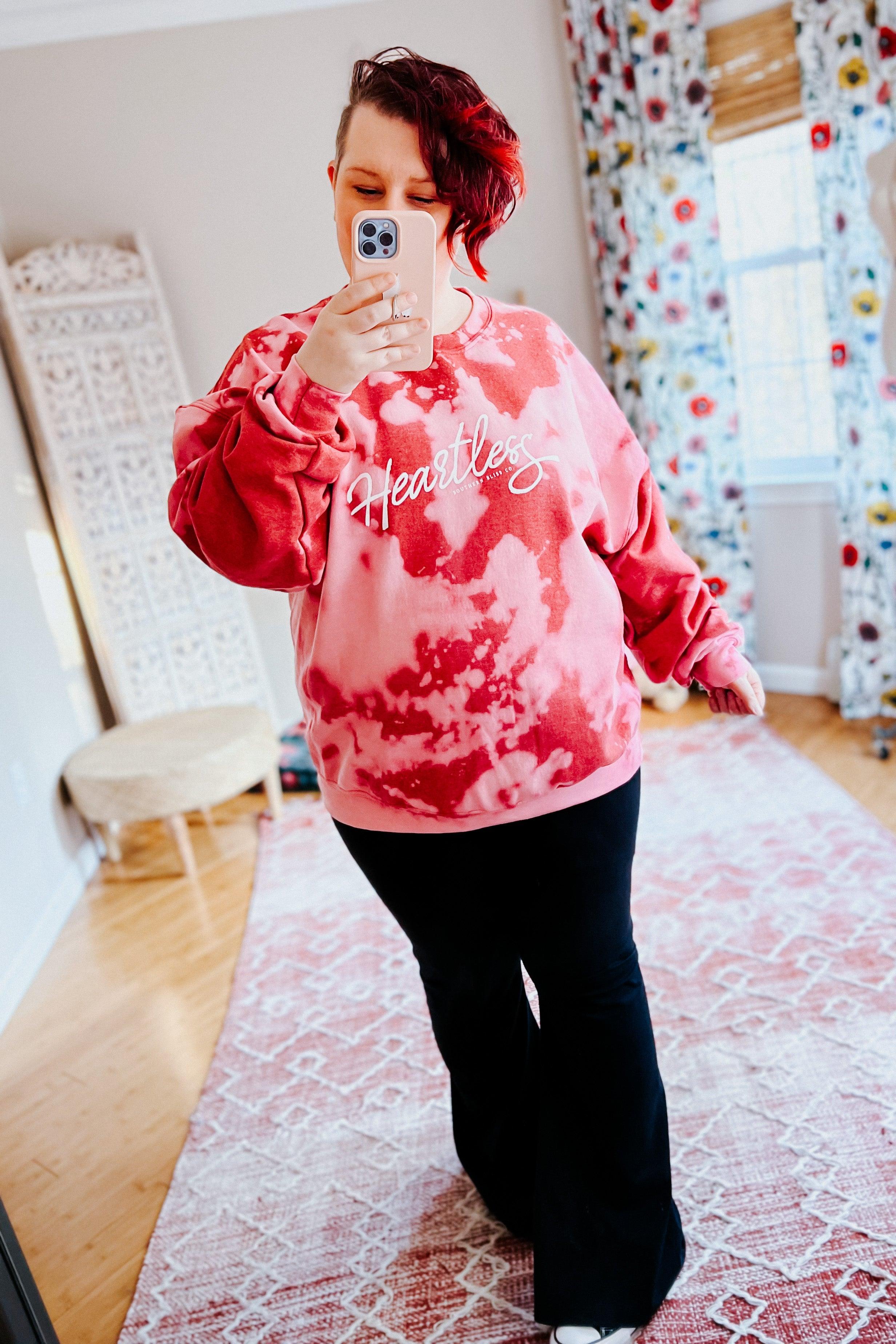 Woman taking mirror selfie wearing red heartless bleached pullover with white script logo and black pants indoors