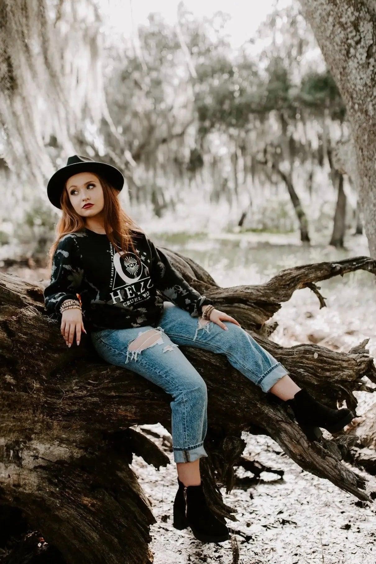 Woman sitting on large tree branch wearing black Hello Cruel World bleached sweatshirt, ripped jeans, black ankle boots, and hat outdoors