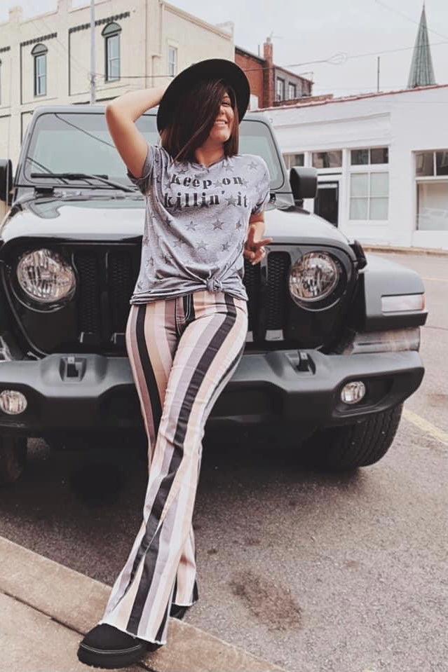 Woman standing outdoors wearing grey Keep On Killin’ It Star Tee tied at the waist with black hat and striped pants