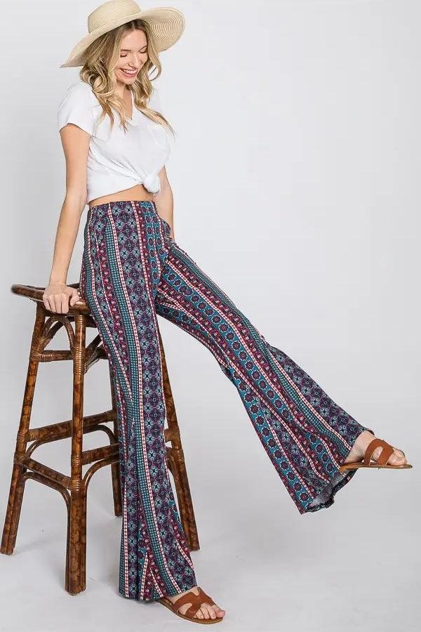 Woman wearing blue patterned flare pants with a white knotted top and straw hat sitting on a wooden stool
