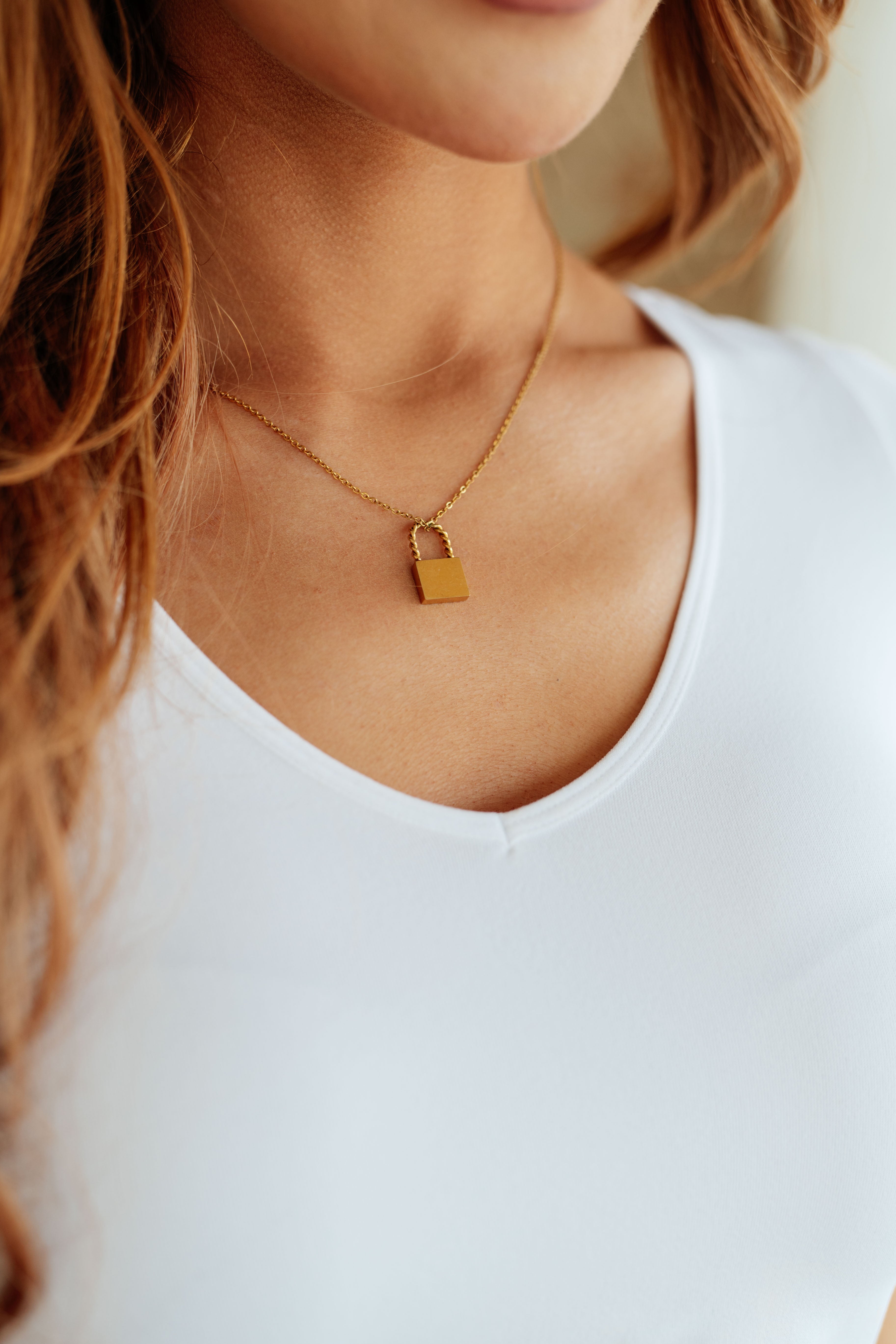 Close-up of a woman wearing a gold padlock pendant necklace on a delicate chain with a white top visible