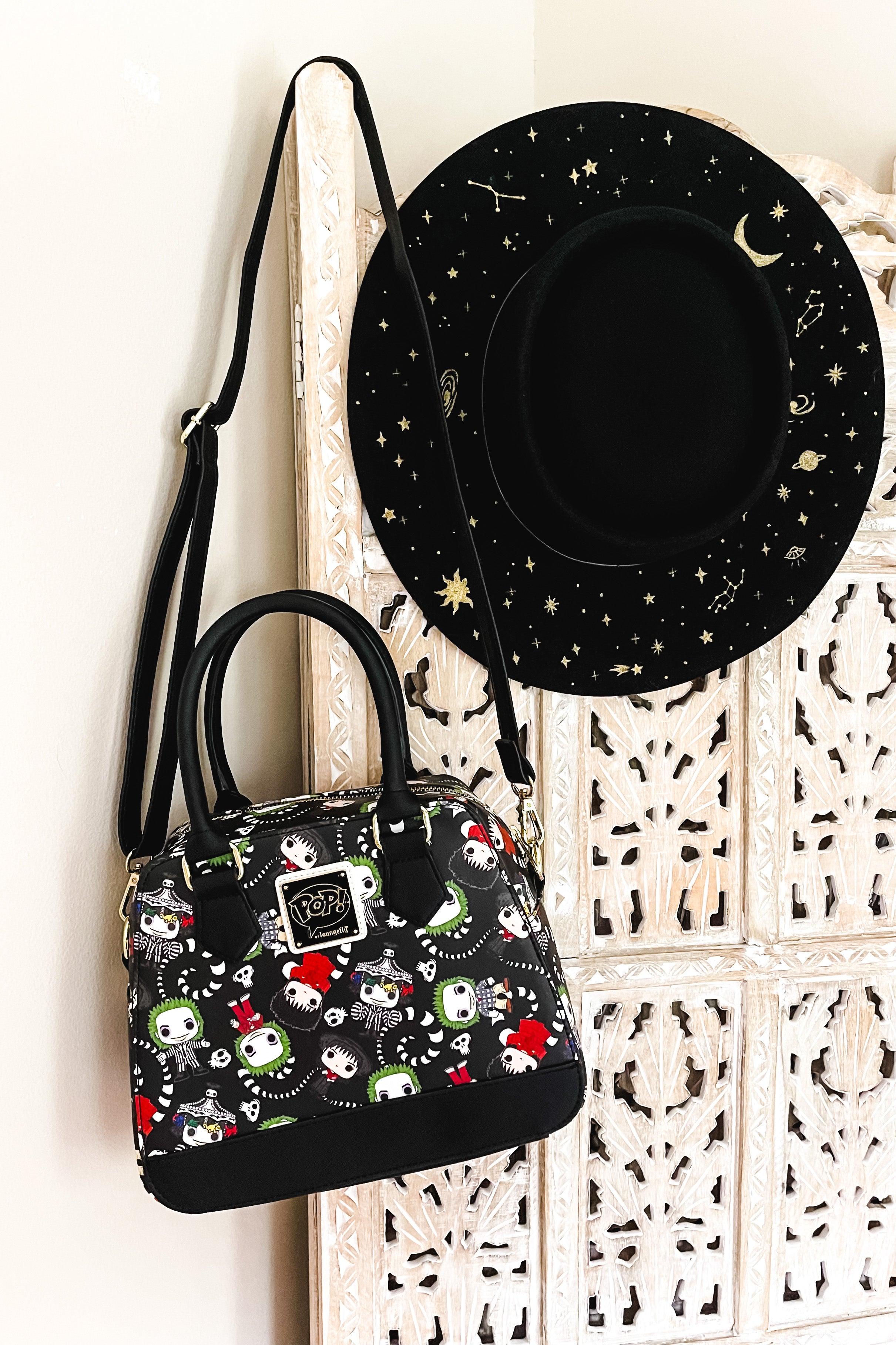 Black loungefly handbag with colorful beetlejuice characters and skulls pattern hanging next to a black wide-brim hat with gold celestial designs