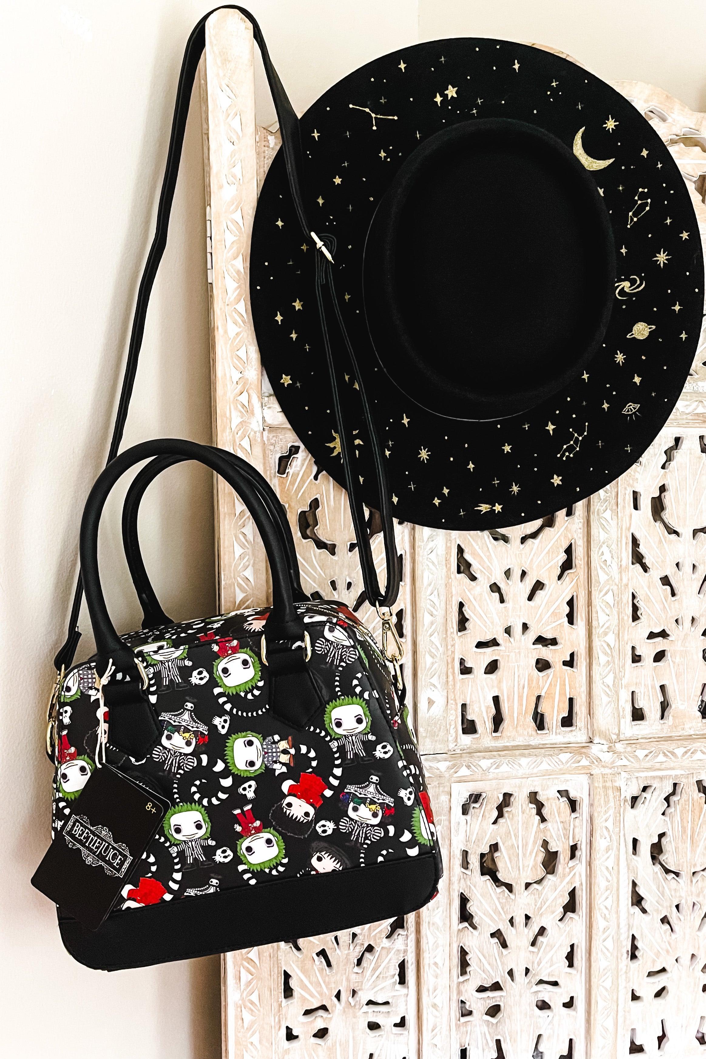 Black loungefly handbag with colorful beetlejuice skeleton characters and red accents hanging beside black wide-brim hat with gold celestial pattern