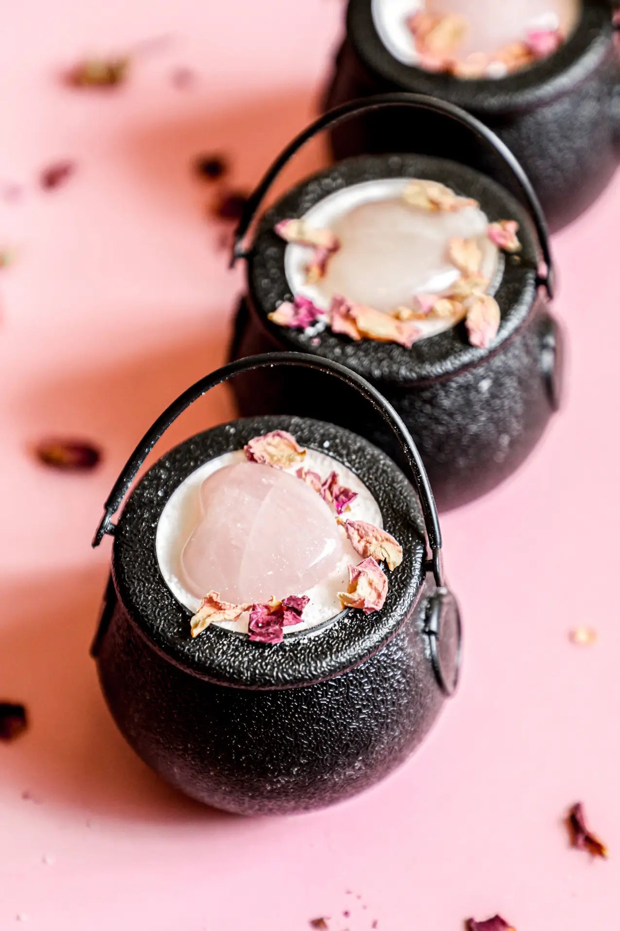 Small black cauldron bath bombs, topped with pink rose quartz hearts and dried flower petals on pink background