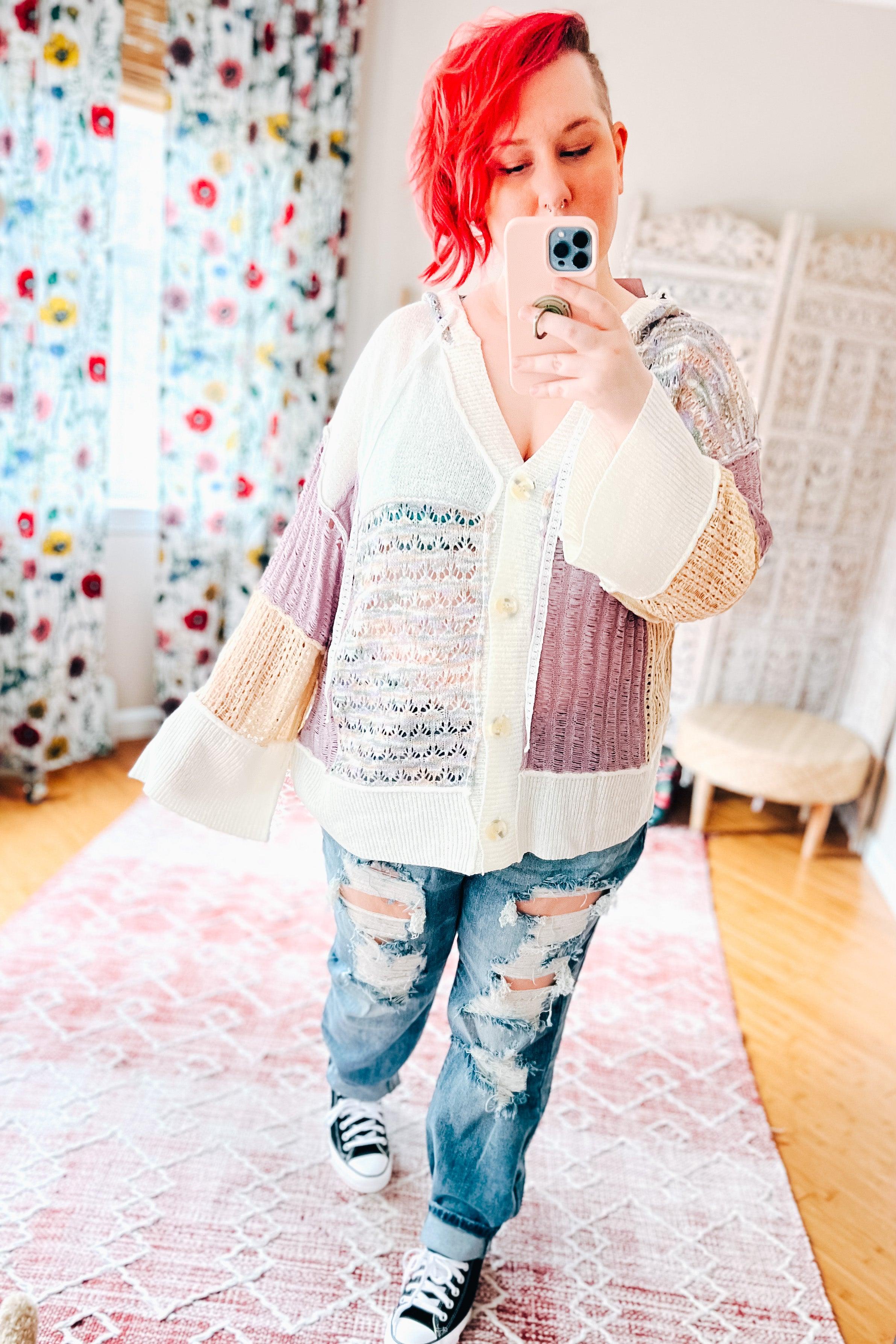 Woman wearing oversized patchwork cardigan with lavender, cream, and beige knit panels paired with ripped jeans and sneakers indoors