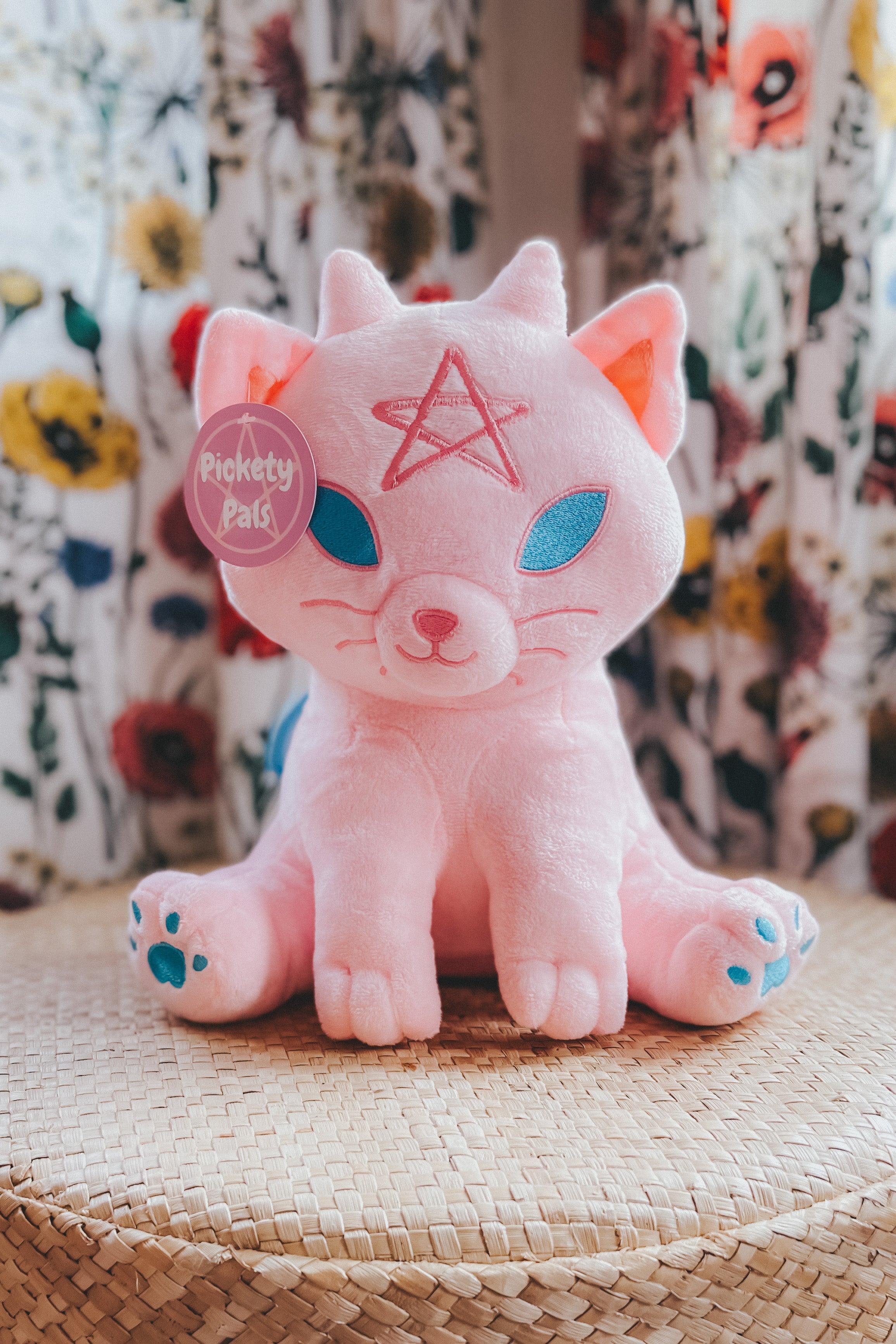 Plush pink cat with embroidered blue eyes and paws and a pentagram on its forehead sitting on woven surface against floral backdrop