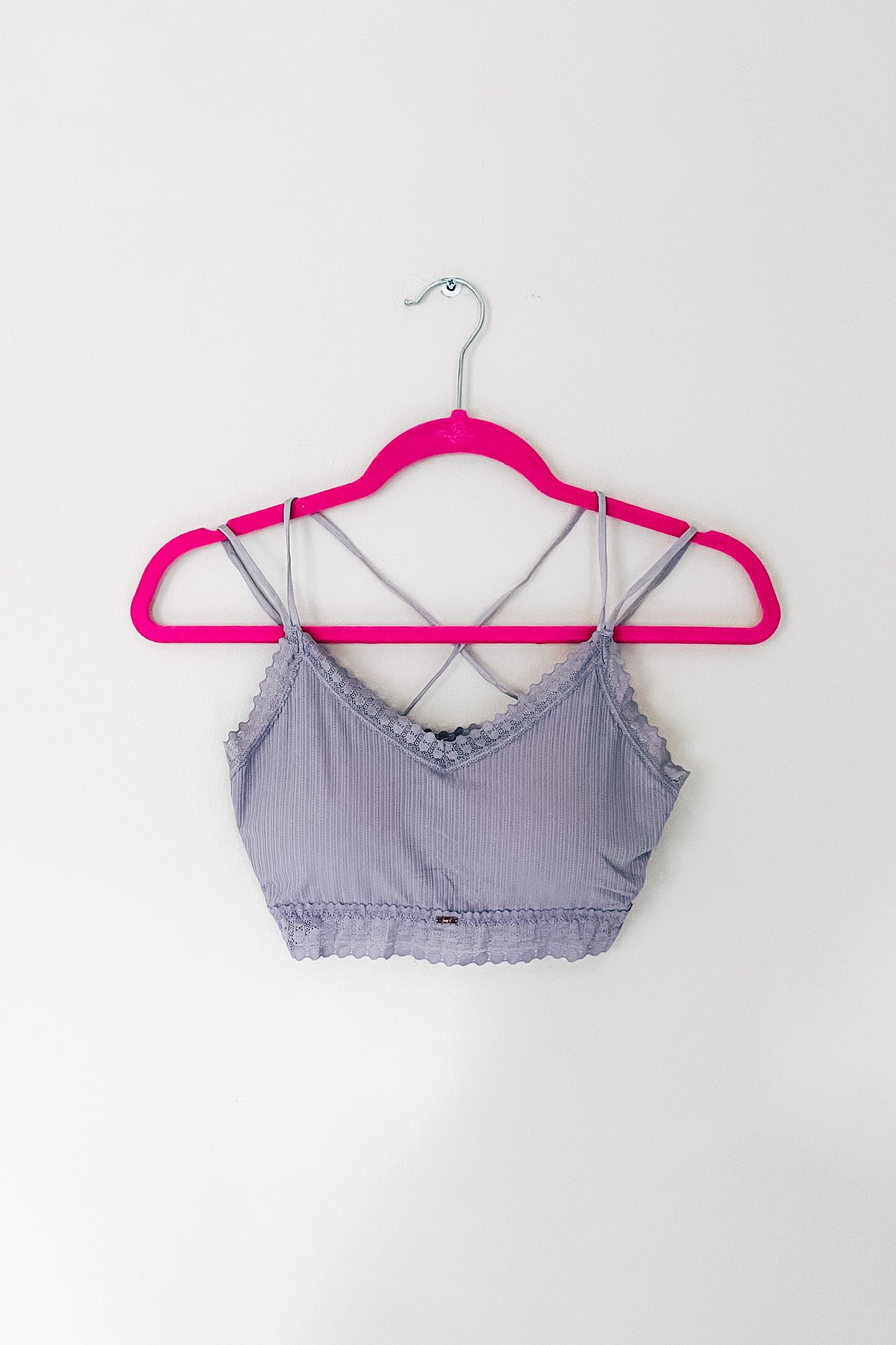 Lilac ribbed bralette with lace trim and crisscross straps displayed on a bright pink hanger against white wall