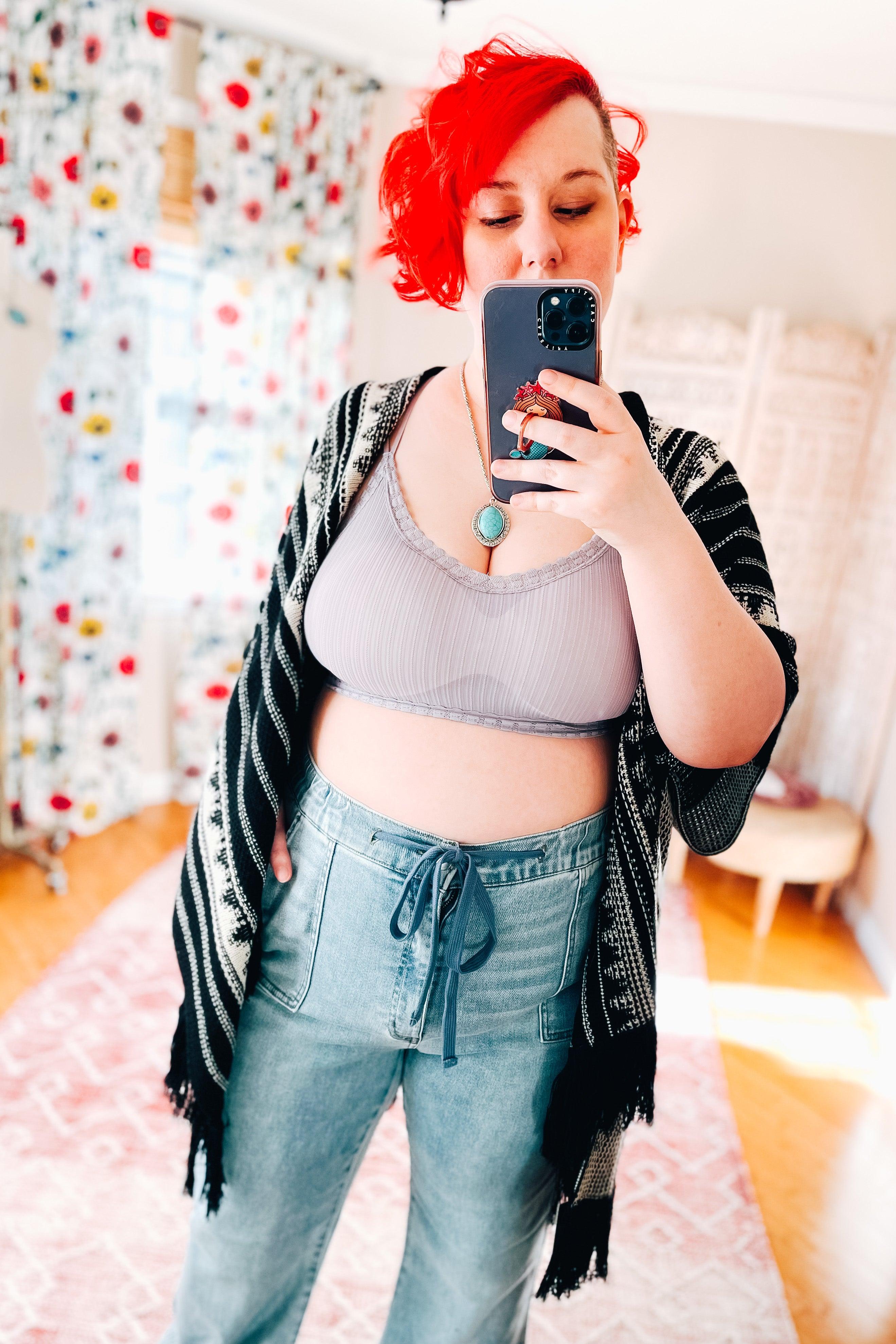 Woman with red hair wearing a lilac ribbed bralette with lace trim, high-waisted denim pants, and a black patterned shawl taking a selfie